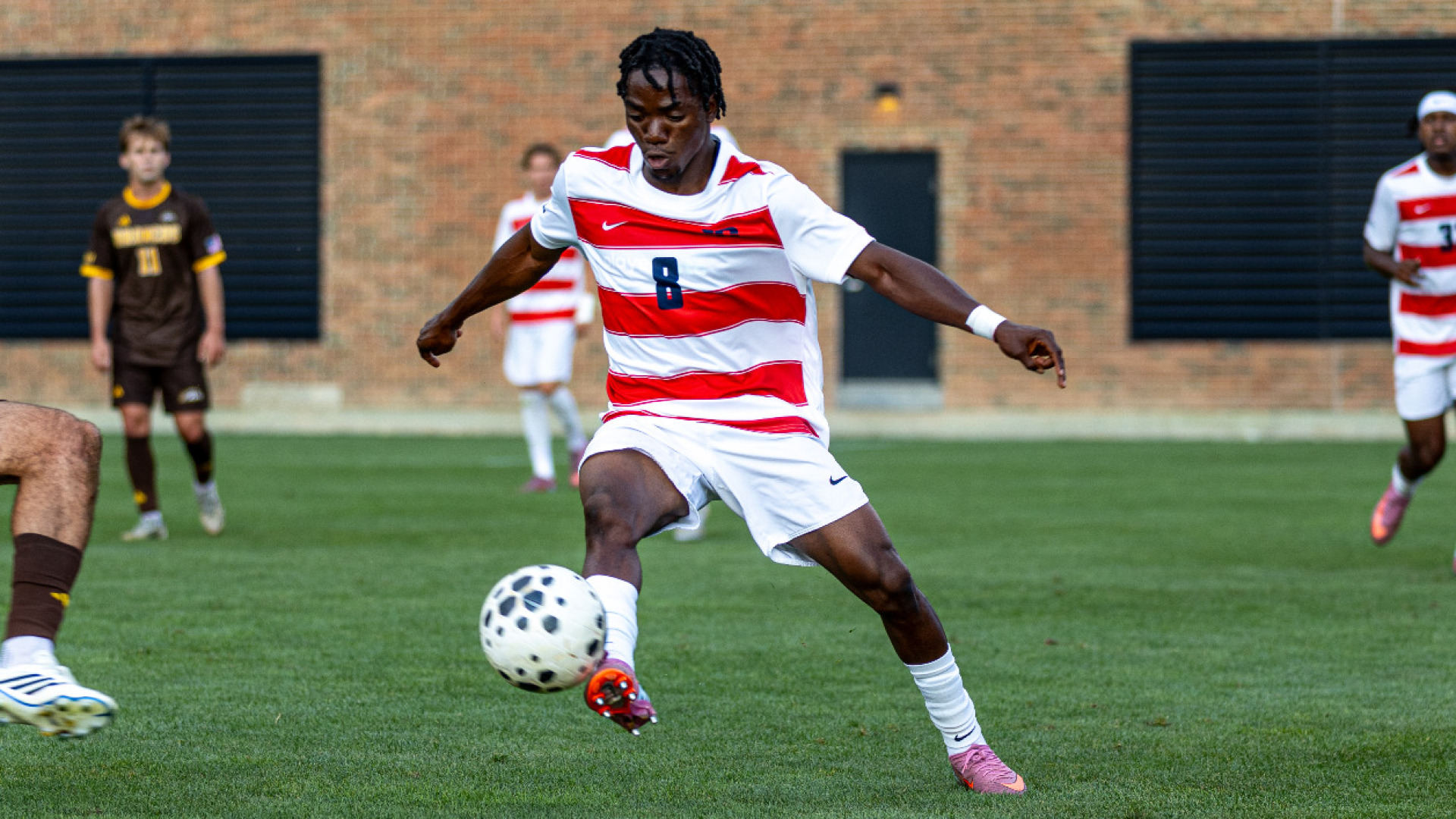 Men's soccer player Farouq Ogundare, wearing a red and white jersey with the number eight with white shorts and white socks, dribbles a soccer ball away from a defender during a game at Baujan Field