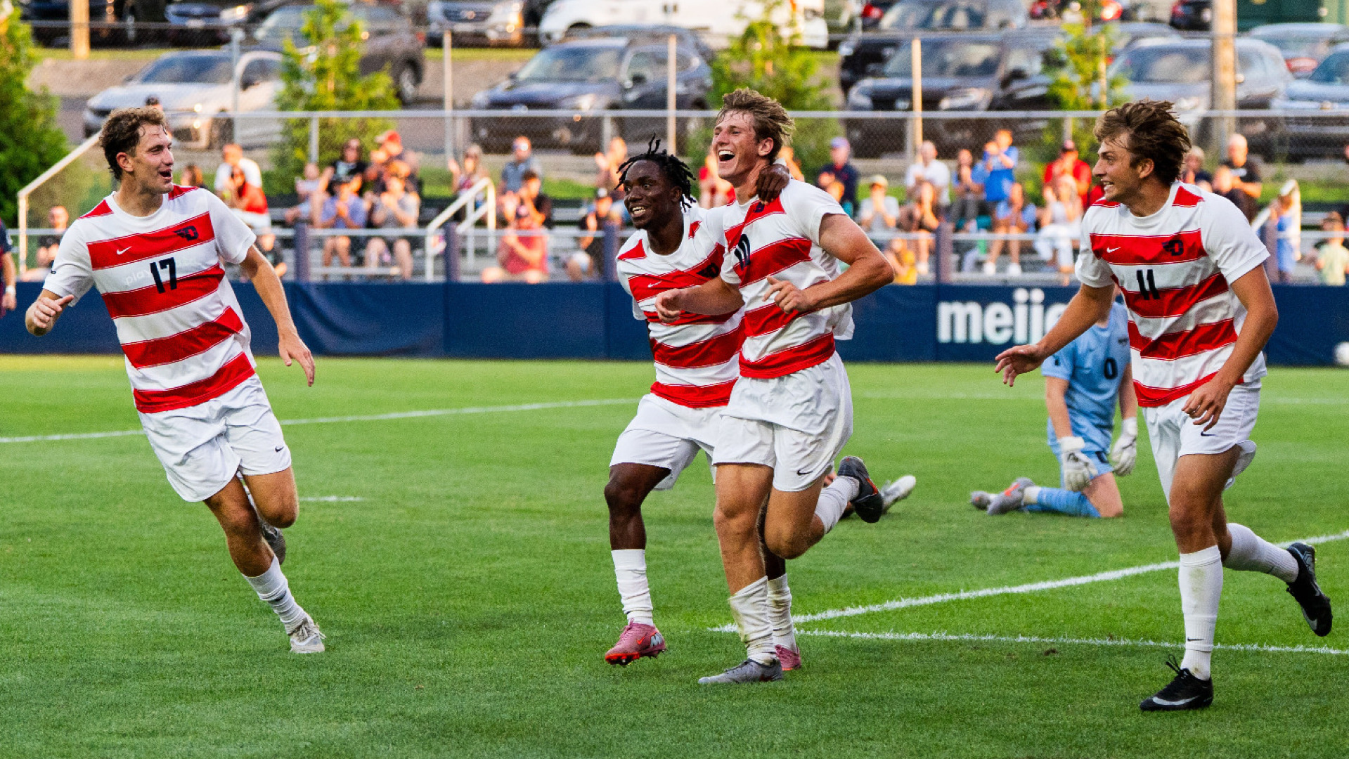Members of the men's soccer team, wearing red and white jerseys with white shorts and socks, celebrate following a goal at Baujan Field