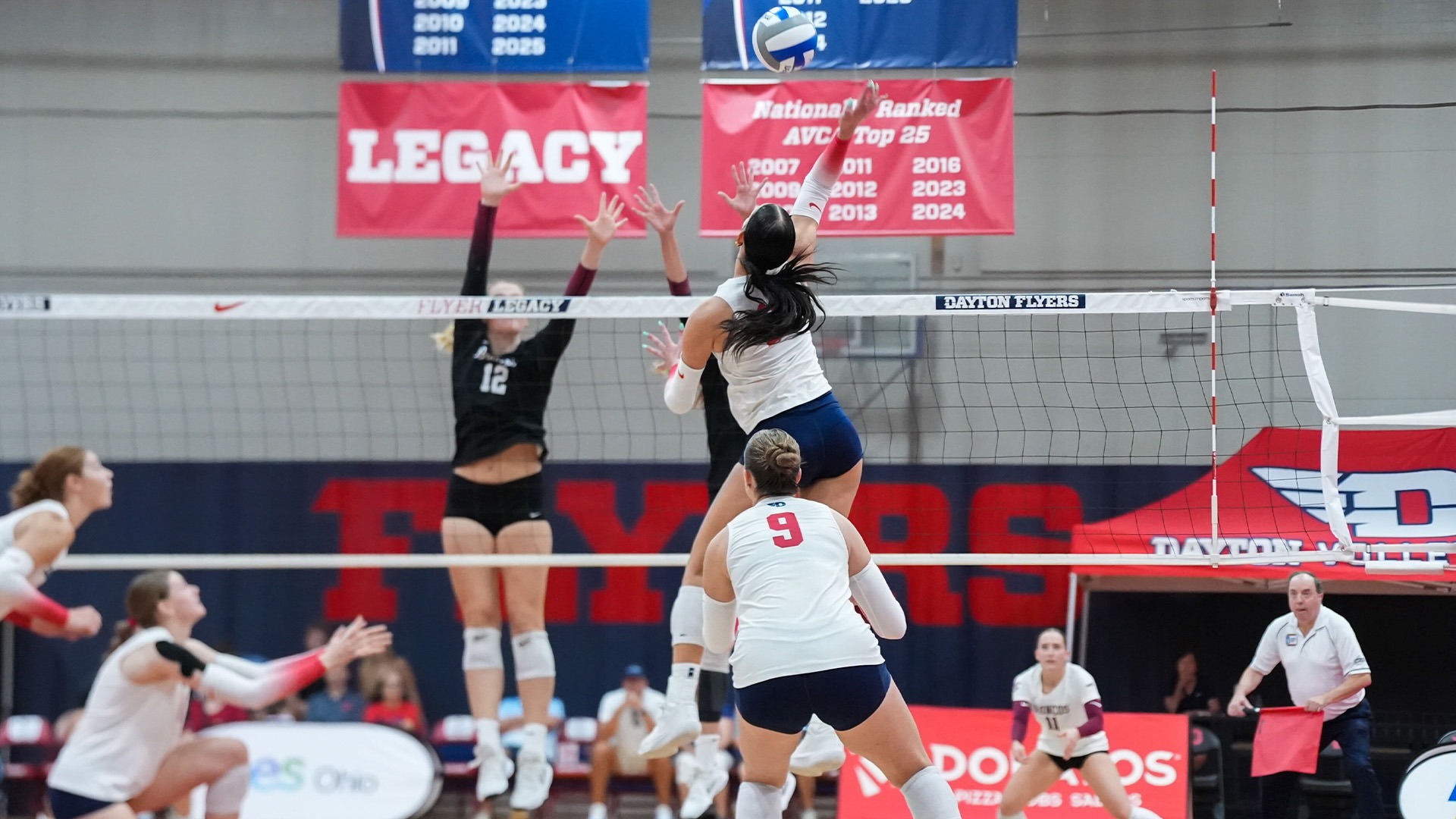 Charlotte Vinson is in the air attack the ball, with Dayton players surrounding her to anticipate a block. The team is in white jerseys with red numbers and navy shorts.