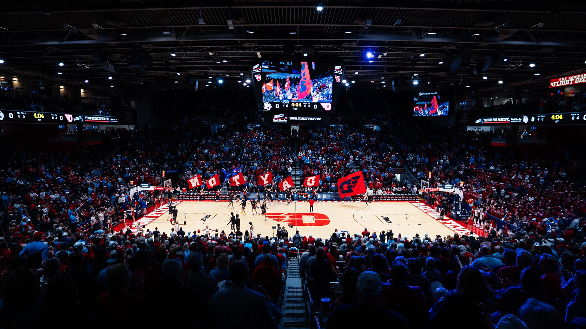 A wide-angle view of a packed basketball arena before tipoff at the University of Dayton. Fans fill the stands as members of the spirit squad run across the court waving large red flags that spell “DAYTON” alongside a Flyers logo flag. Players warm up on the court beneath a center-hung video board displaying pregame festivities, while the brightly lit hardwood contrasts with the dimly lit crowd, creating an energetic game-day atmosphere.