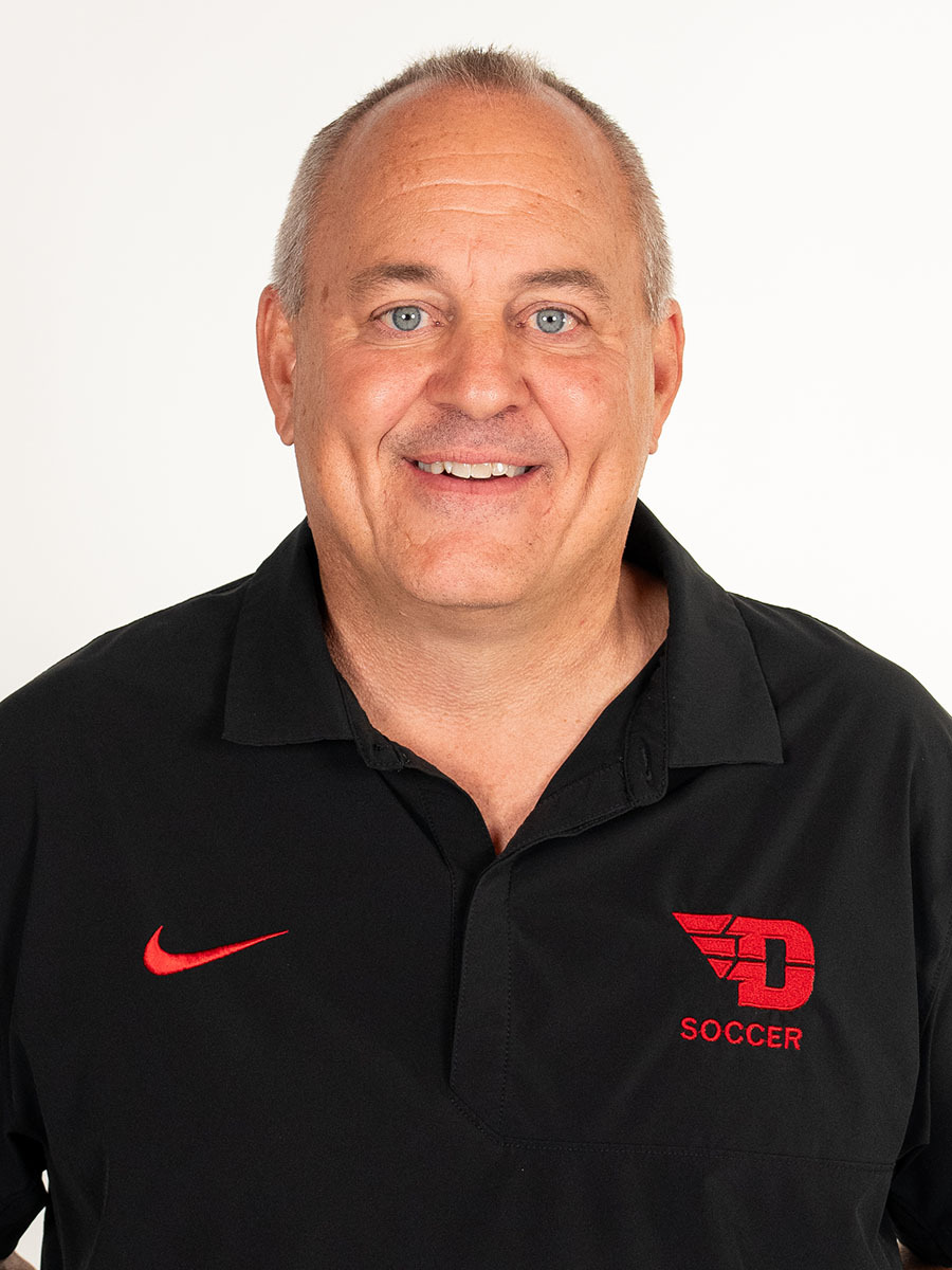 Portrait image of men's soccer head coach Dennis Currier wearing a black polo-style shirt with a red flying D logo