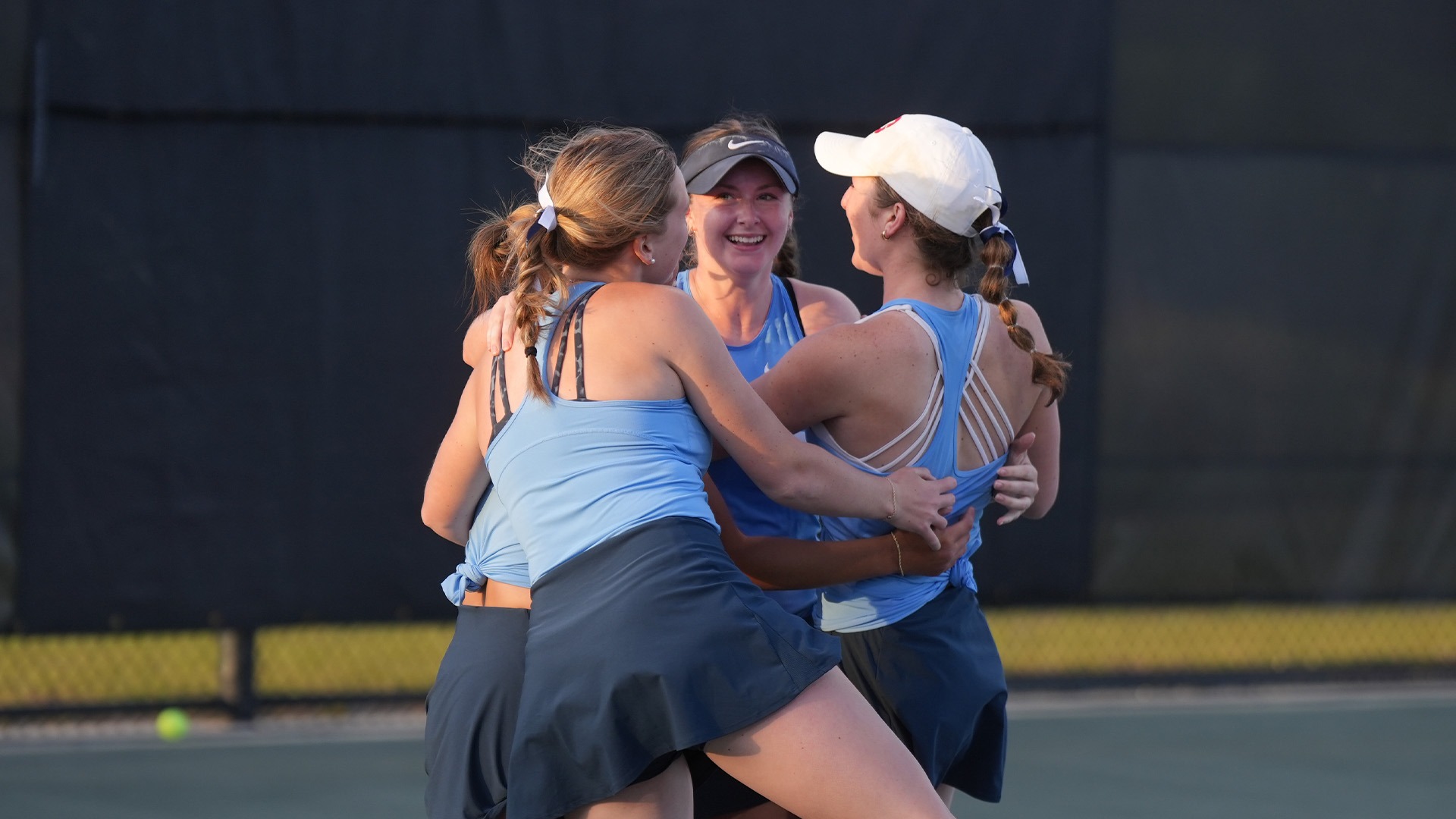 Women's tennis players, wearing Chapel Blue tank tops and navy skirts, celebrate after winning the match.