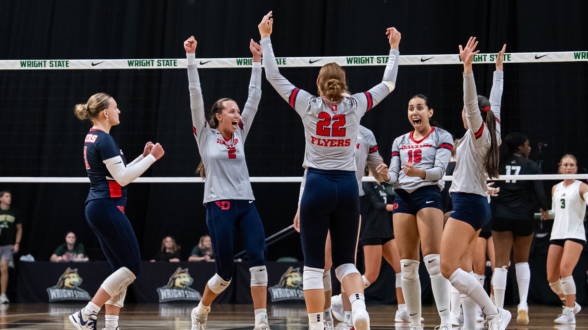 Volleyball players have their arms up in the air in celebration after a point. They are wearing grey jerseys and navy pants. The libero on the left is wearing all navy.