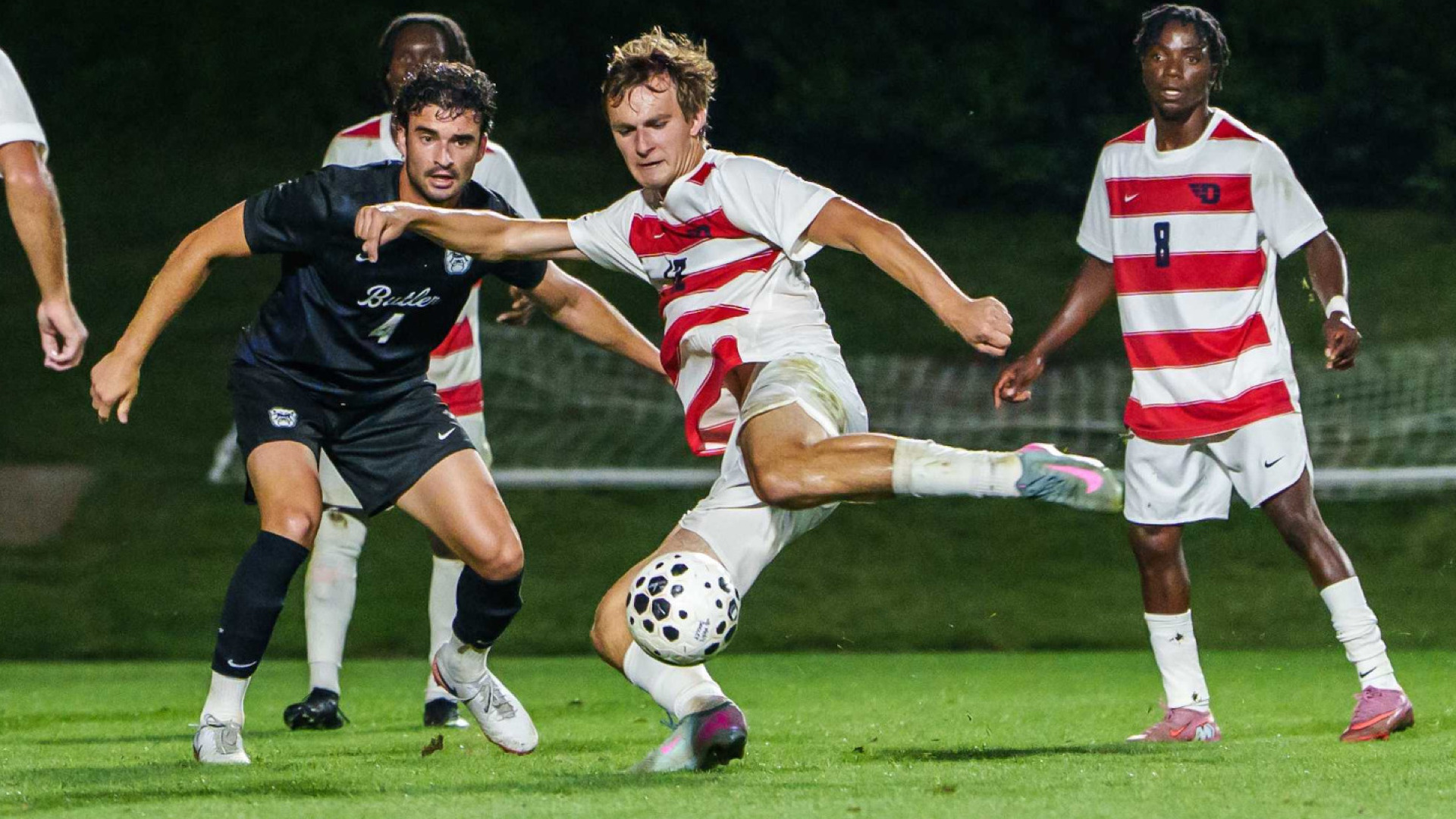 Men's soccer player Cooper Lijewski, wearing a red and white striped jersey with the number 12 along with white shorts and white socks, shoot the ball during a game at Baujan Field against Butler
