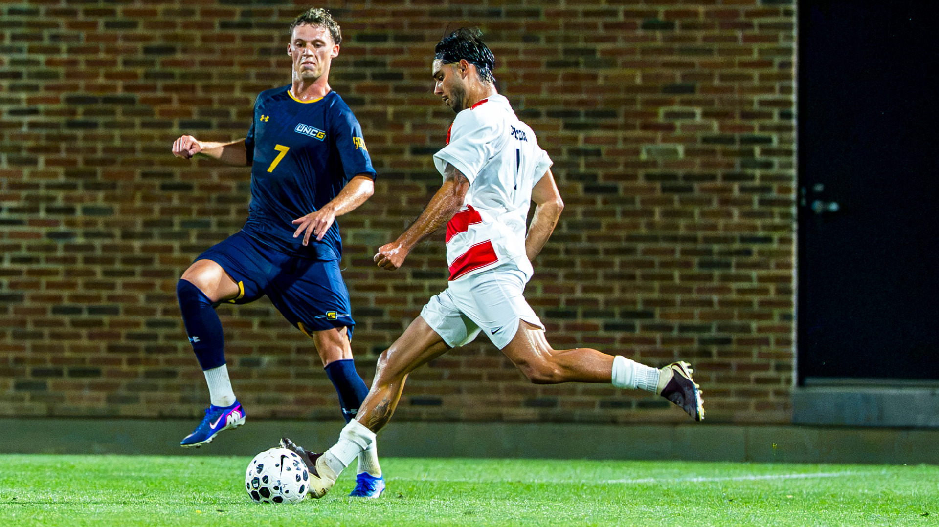 Men's soccer player Pablo Martinez, wearing a red and white striped jersey and white shorts, prepares to kick a ball past a defender wearing a blue uniform during a match at Baujan Field