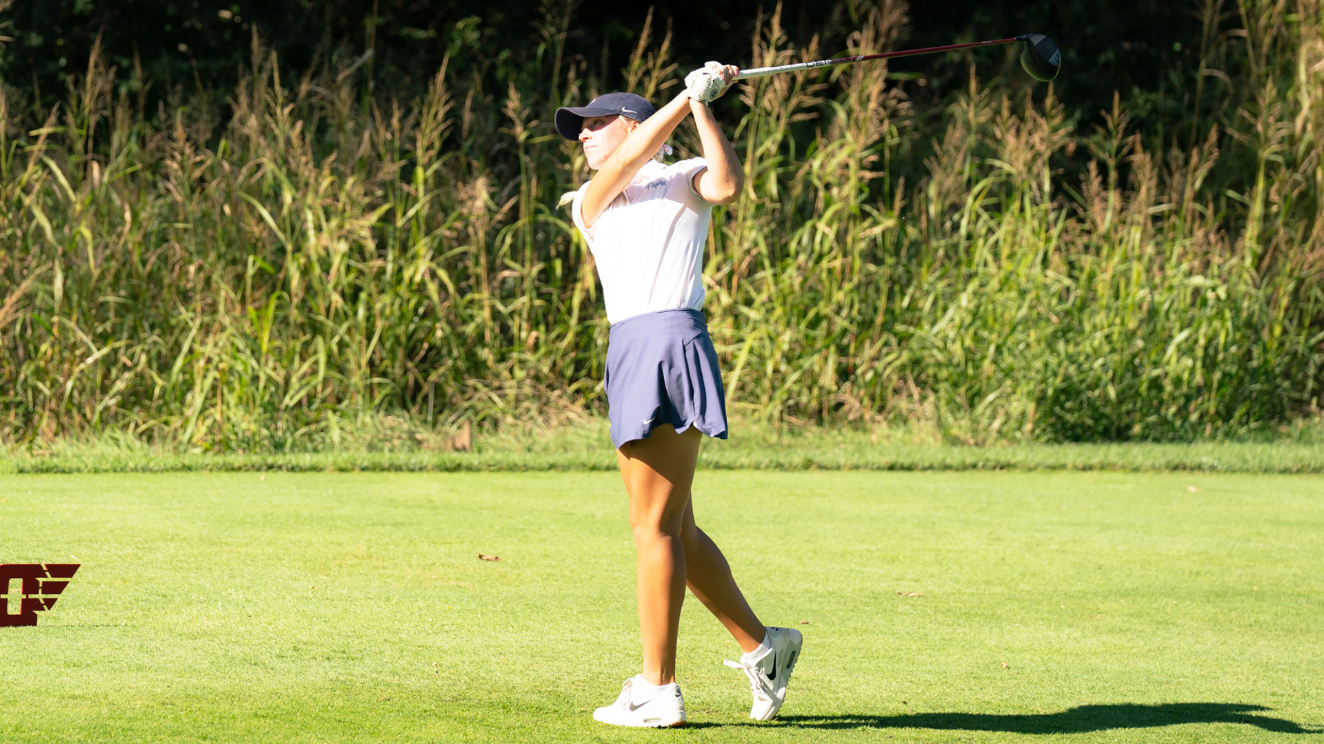 Women's golfer Kylee Heidemann, wearing a white polo-style shirt, blue skirt and blue hat, at the top of her back swing in a tee box at NCR Country Club during the Flyer Invitational
