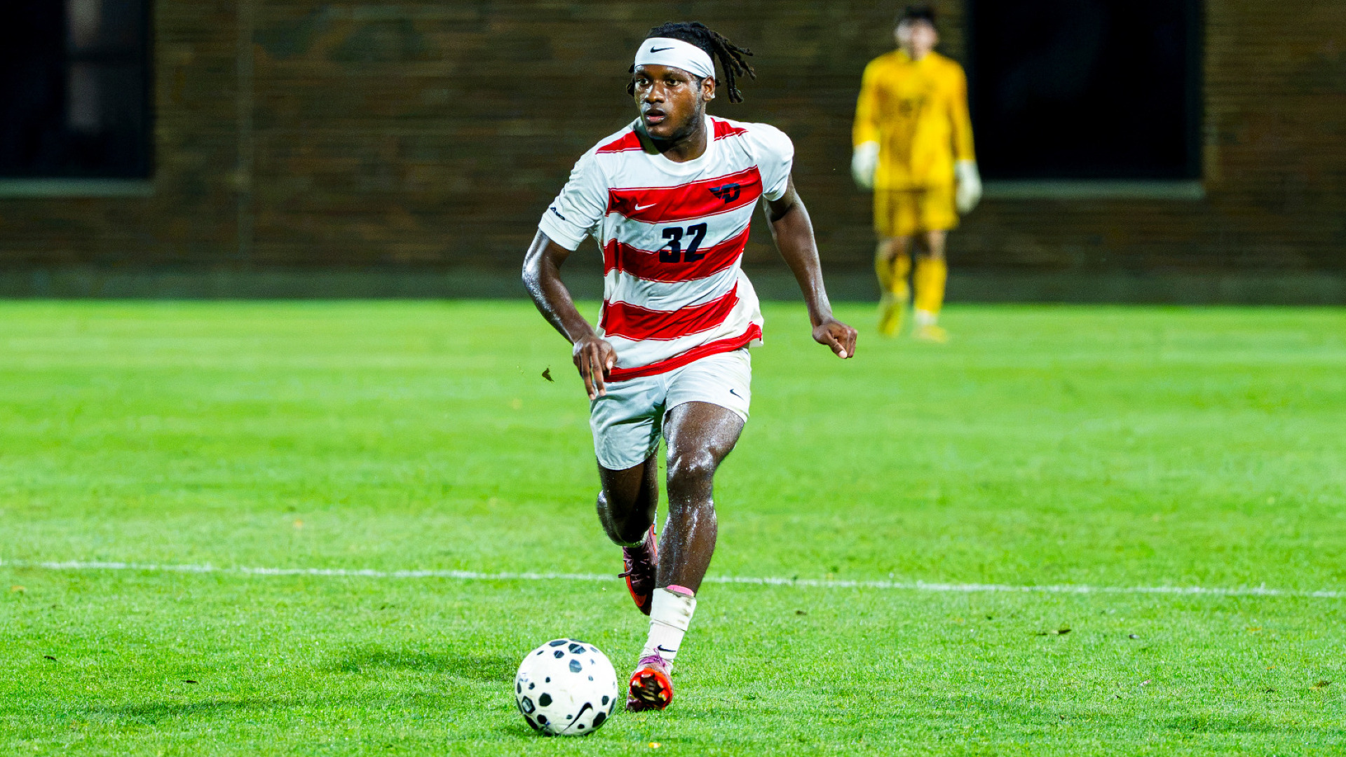 Men's soccer player Chase Jackson, wearing a red and white striped jersey with the number 32 along with white shorts and white headband, dribbles the ball down field during a match at Baujan Field
