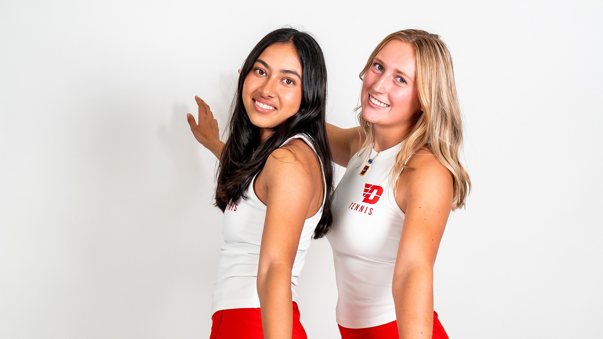 Naima Luthra and Caroline Boyle pose front to back, facing the left. They are wearing white tank tops and red shorts.