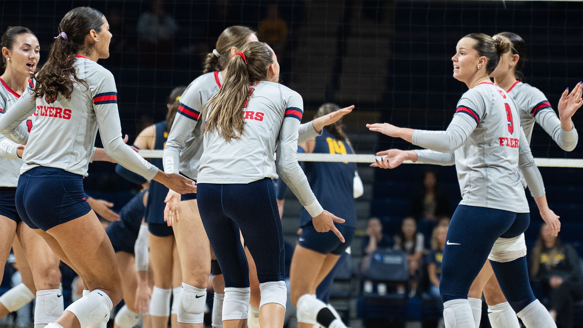Volleyball players on the court high five after a point at Savage Arena. They are wearing grey jerseys with navy shorts and pants.