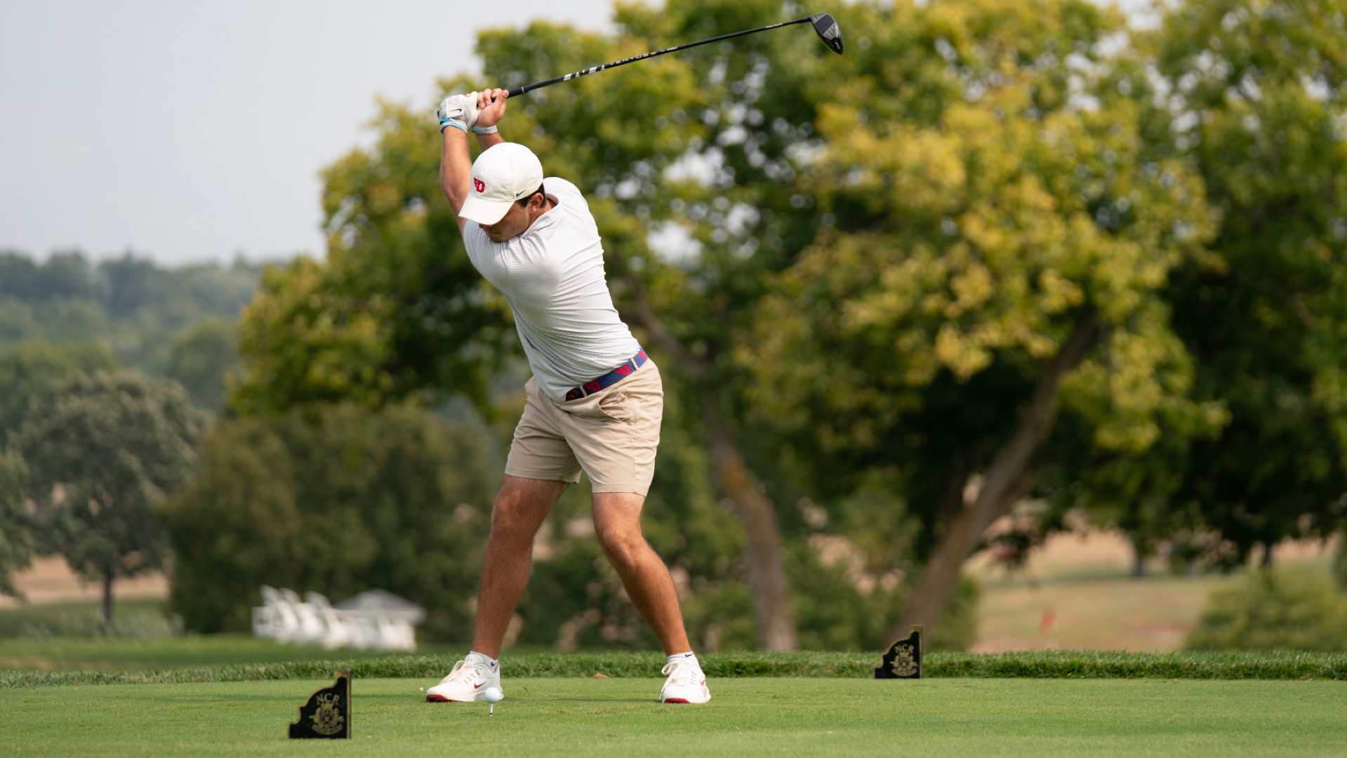Men's golfer Robert Gerwin, wearing a white polo-style shirt, khaki shorts and a white hat, at the top of his back swing in a tee box suring a practice round at NCR Country Club