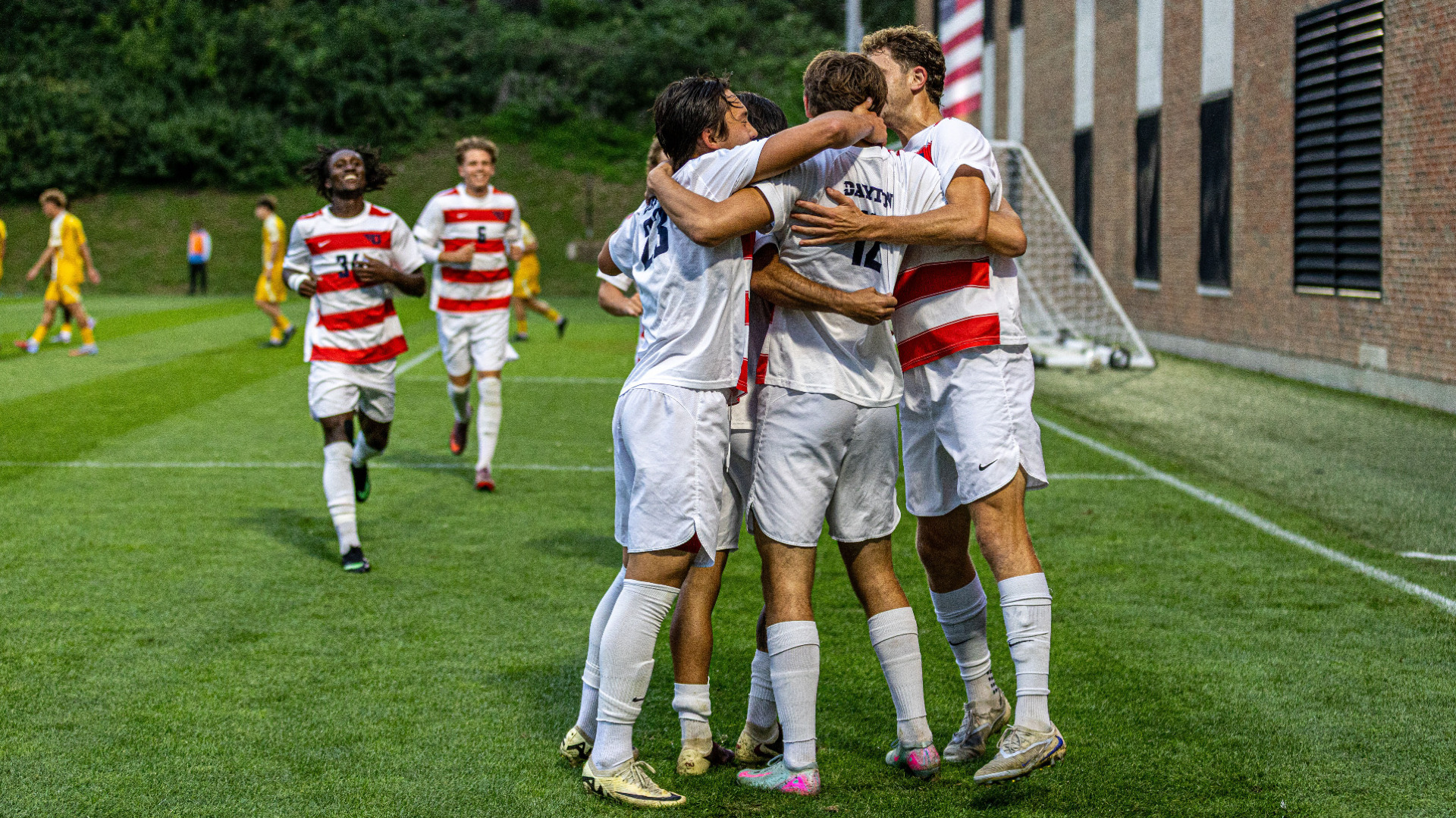 Members of the men's soccer team wearing red and white striped jersey tops with white shorts, embrace and celebrate following a goal by number 12 Cooper Lijewski during a match at Baujan field