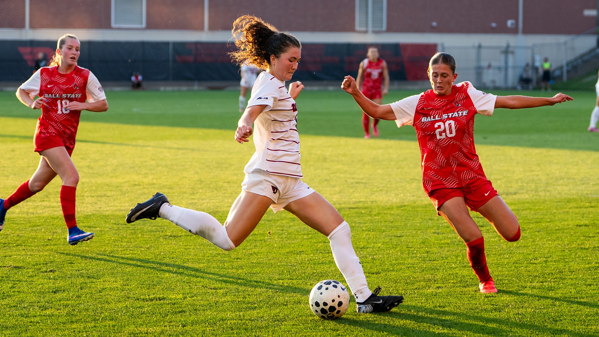 Dayton women’s soccer player Reese Gallagher, in a white uniform, dribbles the ball between two Ball State defenders in red during a match.