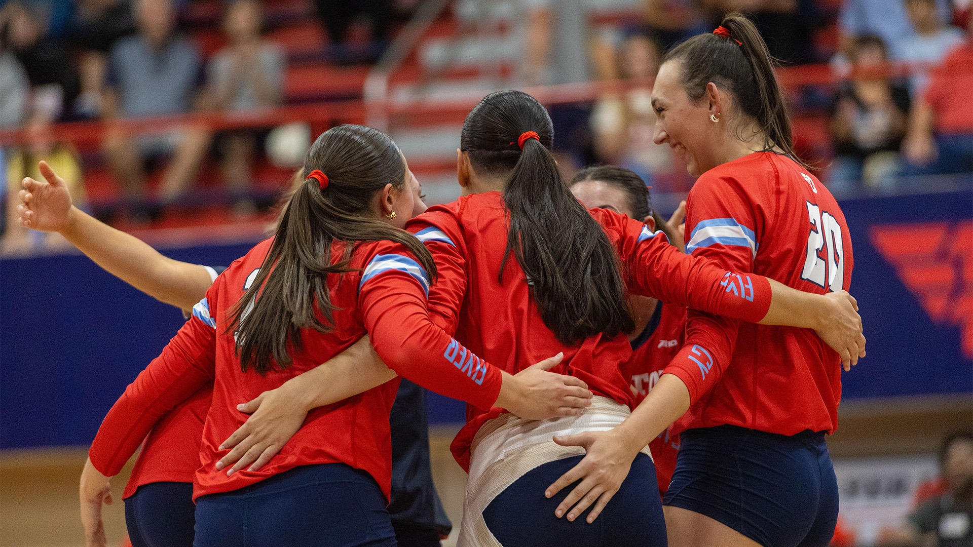 Volleyball players huddle in celebration after a point. They are wearing red jerseys with navy pants and shorts.