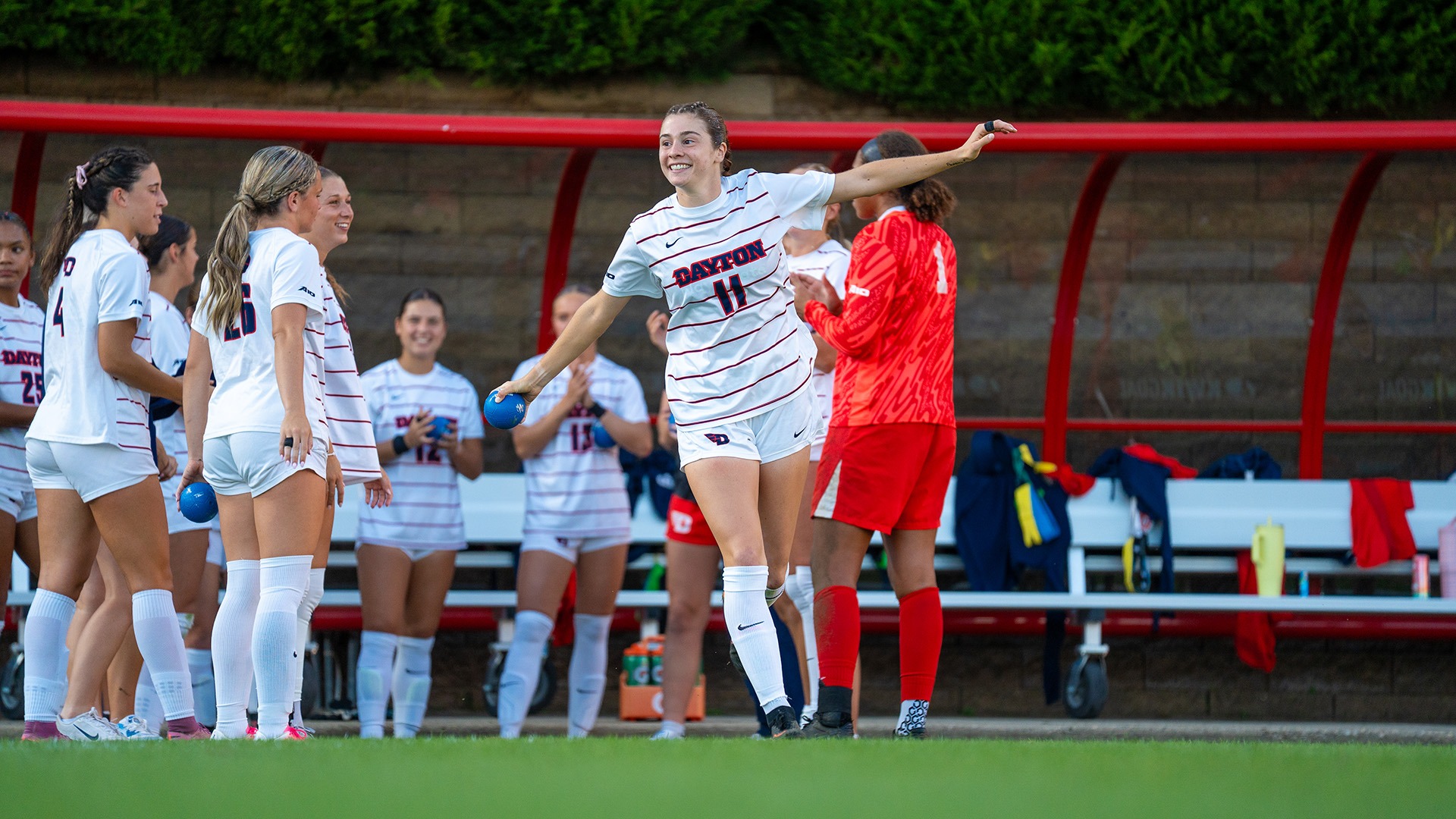 Dayton women's soccer player Liv Grenda, wearing a white jersey with the number 11, runs out pretending to be an airplane.