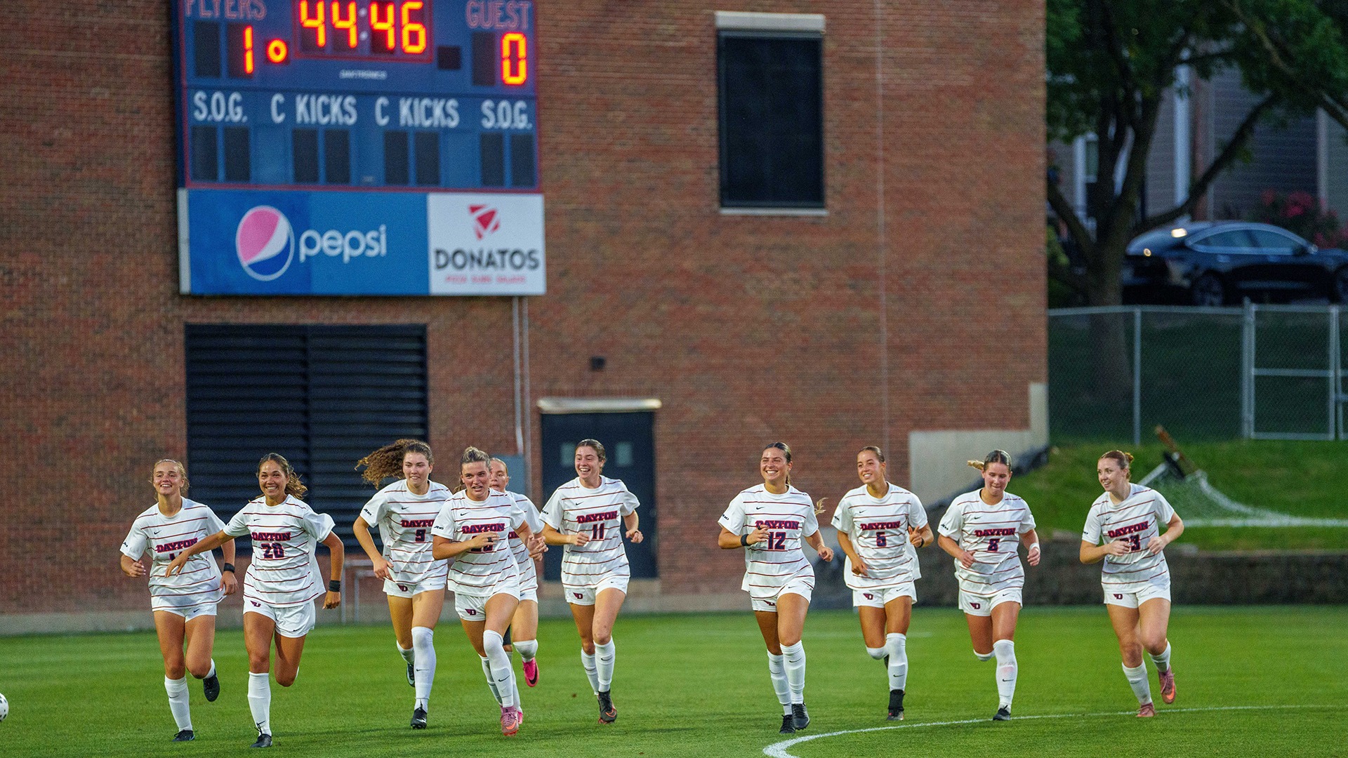 University of Dayton women’s soccer players run together across the field in celebration during a home match, smiling beneath the scoreboard.