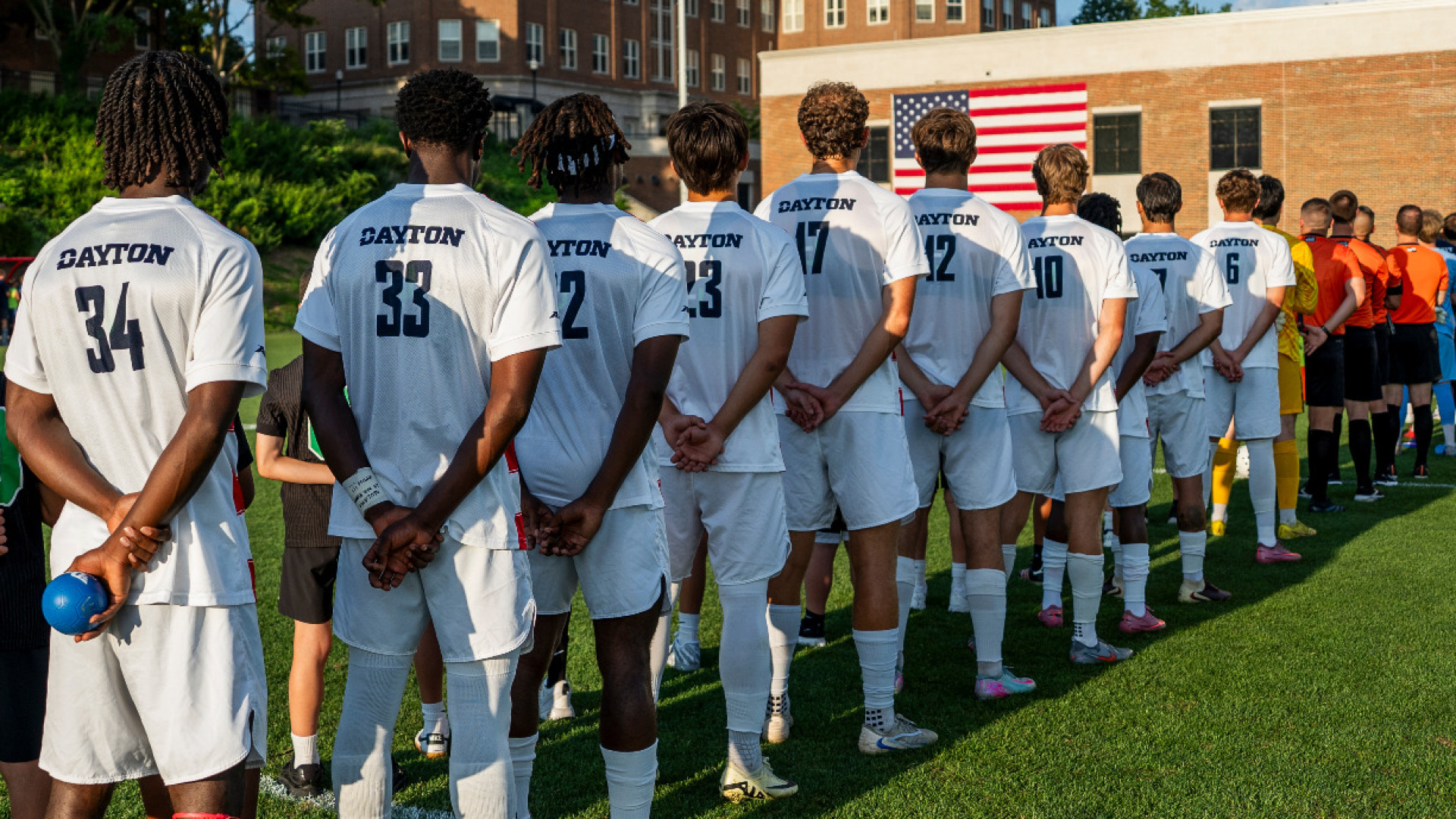 Members of the men's soccer team, wearing white jerseys and white shorts with blue numbers and the Dayton word mark in blue, lined up at middle field with their backs facing the camera during the national anthem at Baujan Field