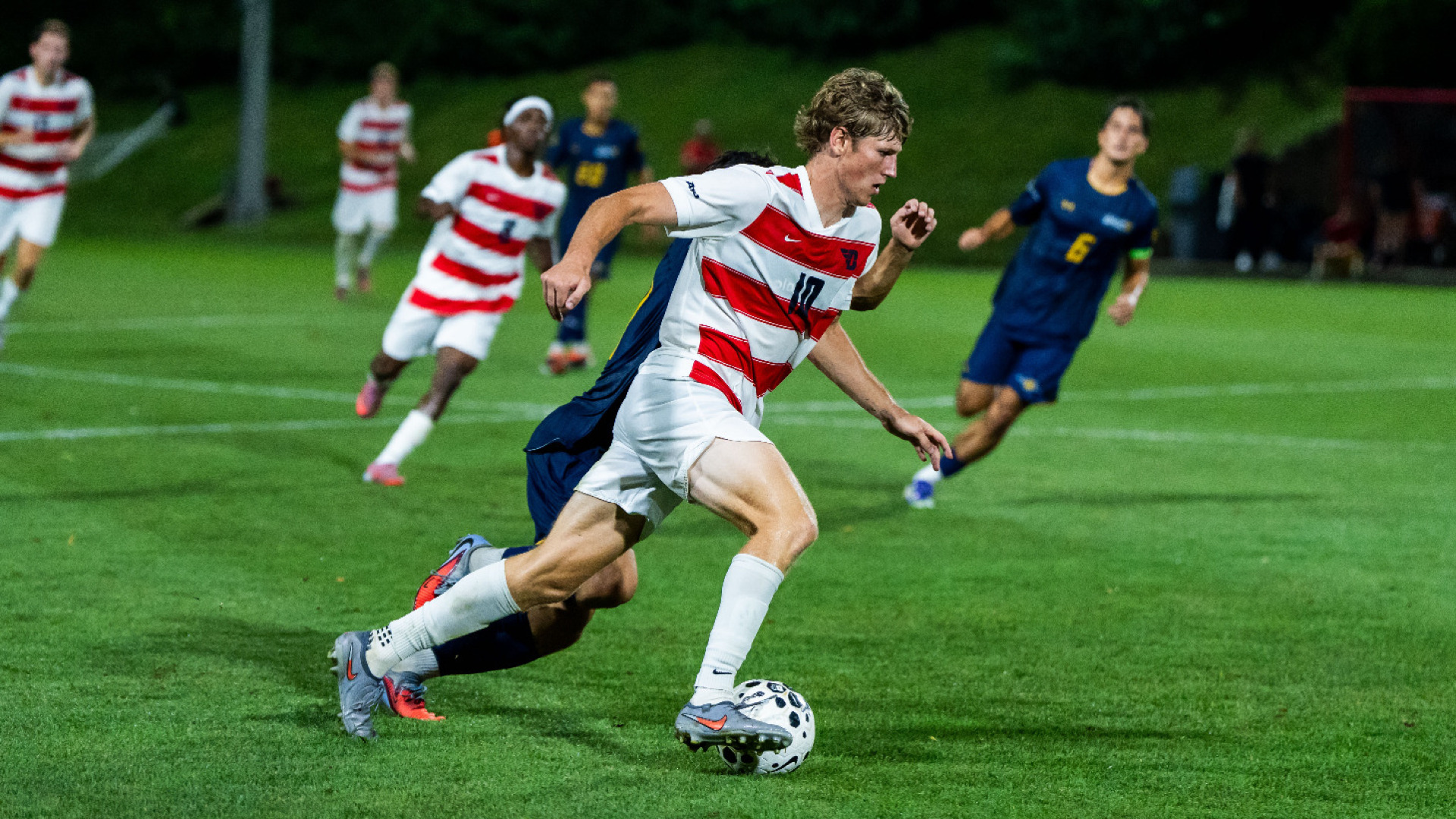 Men's soccer player Braden Seel, wearing a red and white striped jersey, white shorts and white socks, dribbles the ball away from a defender toward the goal during a game at Baujan Field