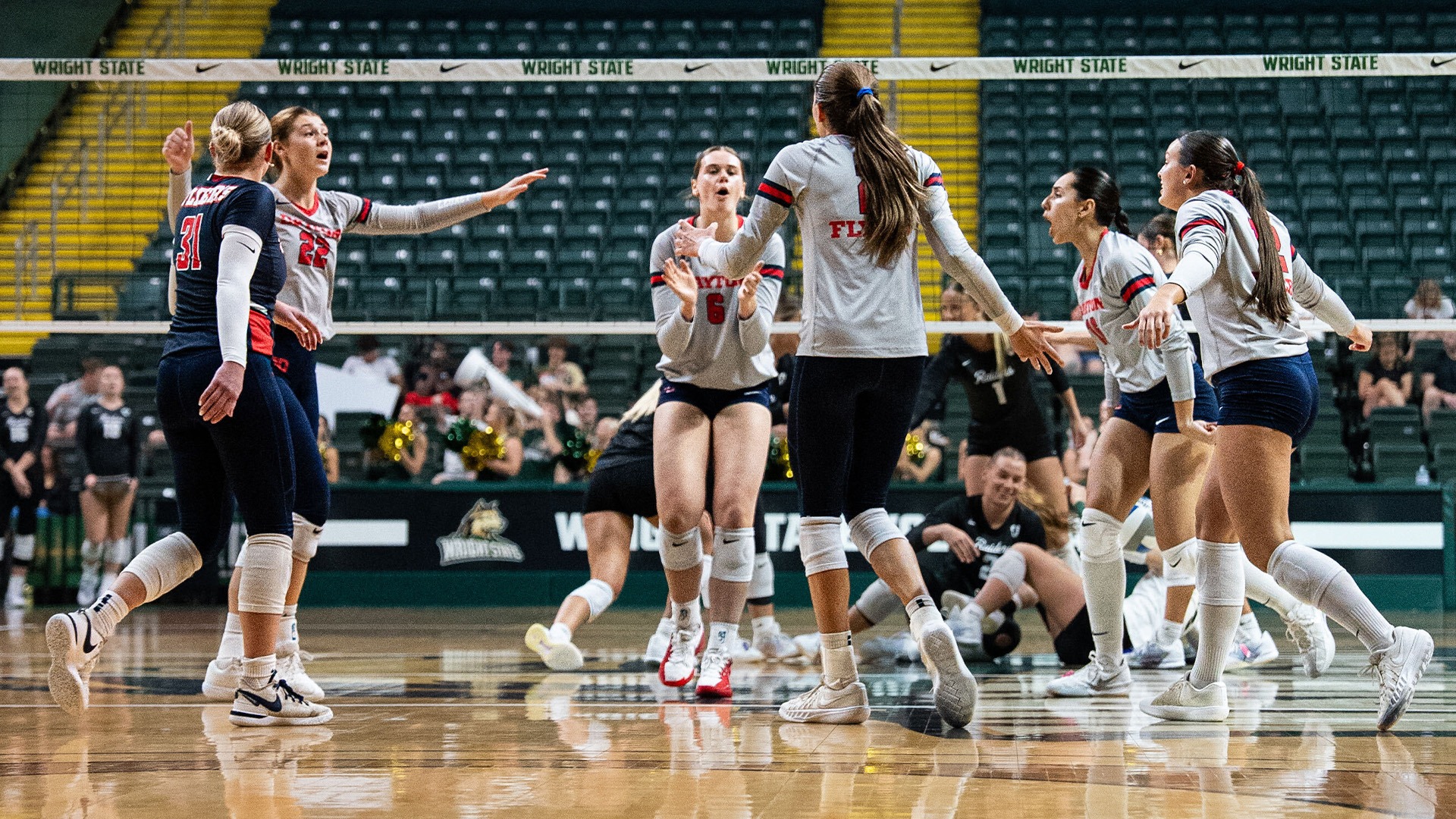 Volleyball players celebrate on the court after a point. They are in gray jerseys and navy bottoms. The libero on the left is wearing all navy.