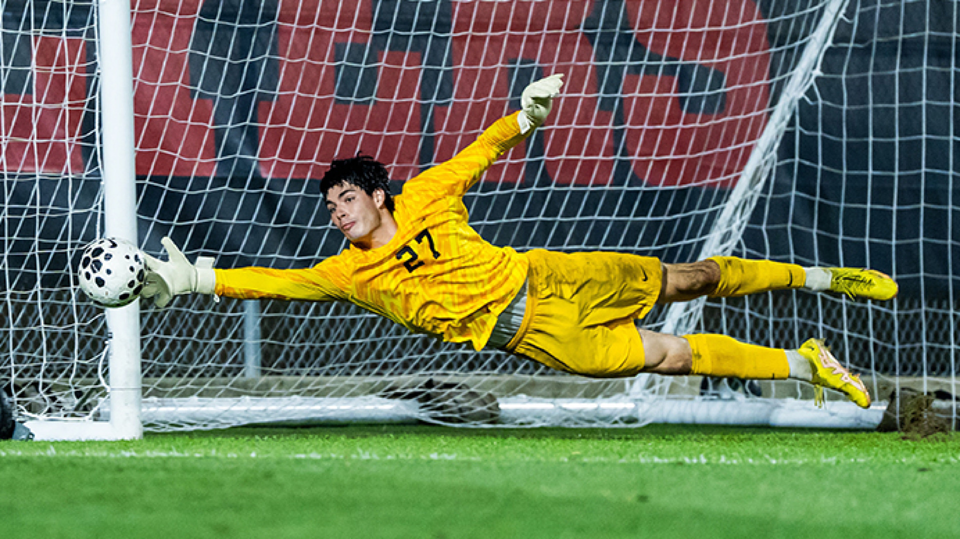 Men's soccer goalkeeper Santino Inkpen, wearing a yellow jersey with the number 27 and yellow shorts, stretches out to save a ball from going into the goal during a match at Baujan Field