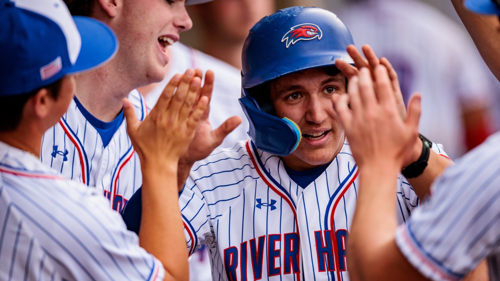 UMass Lowell baseball players in white uniforms high fiving in the dugout after scoring a run 