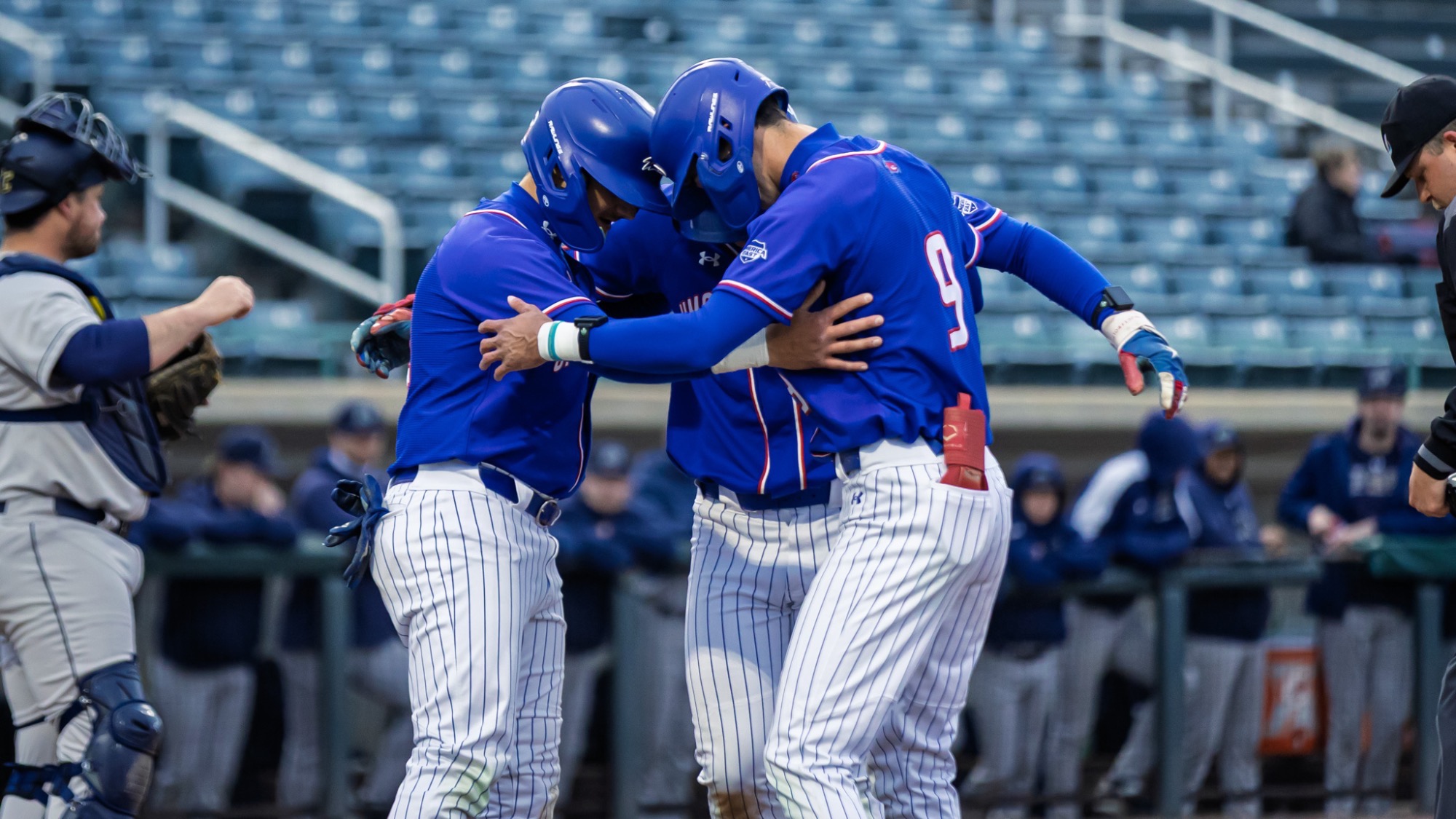 UMass Lowell baseball players in blue uniforms celebrating at home plate after scoring a run in the contest