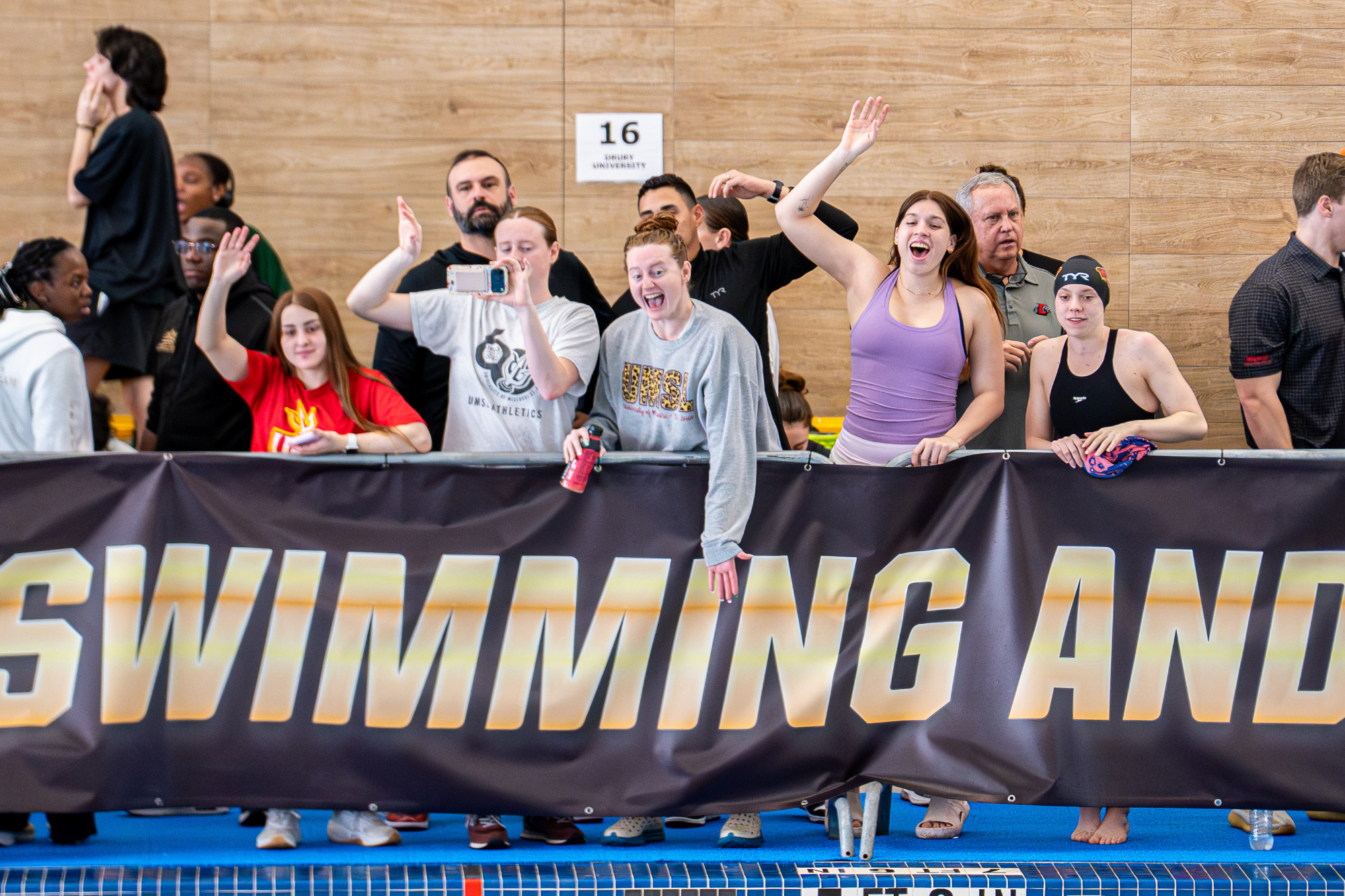 UMSL swimmers (left-to-right) Uxue Ortiz de Pinedo, Ella Lantz, Audrey Lantz, Romina Itzkovich and Justice Beard cheering on Aaron Wicklund in the 400 IM while assistant coach Kale Kirchner and head coach Tony Hernandez watch from behind