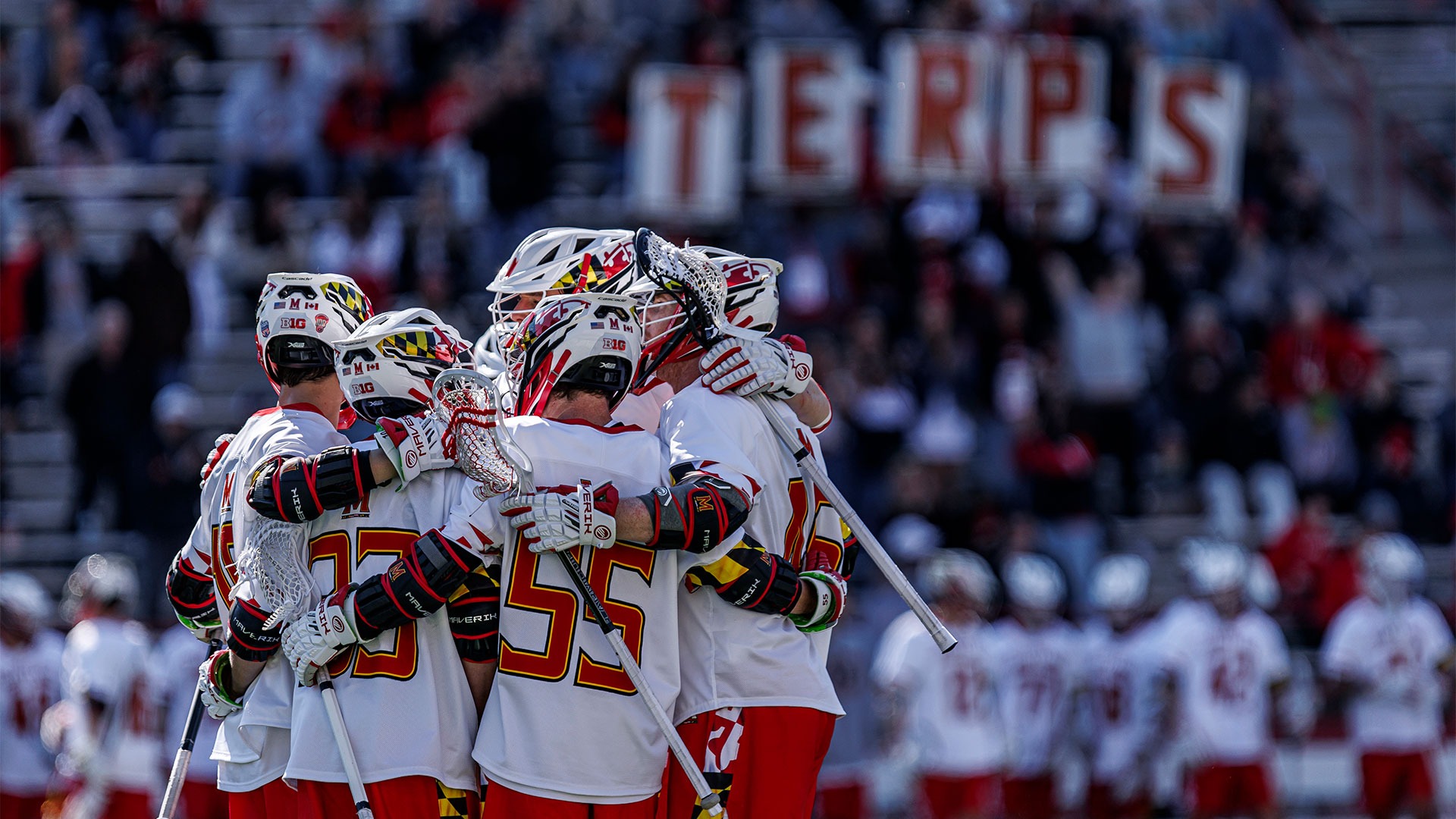 Men's Lacrosse players huddling up at SECU Stadium with fans holding signs that spell out TERPS in the background
