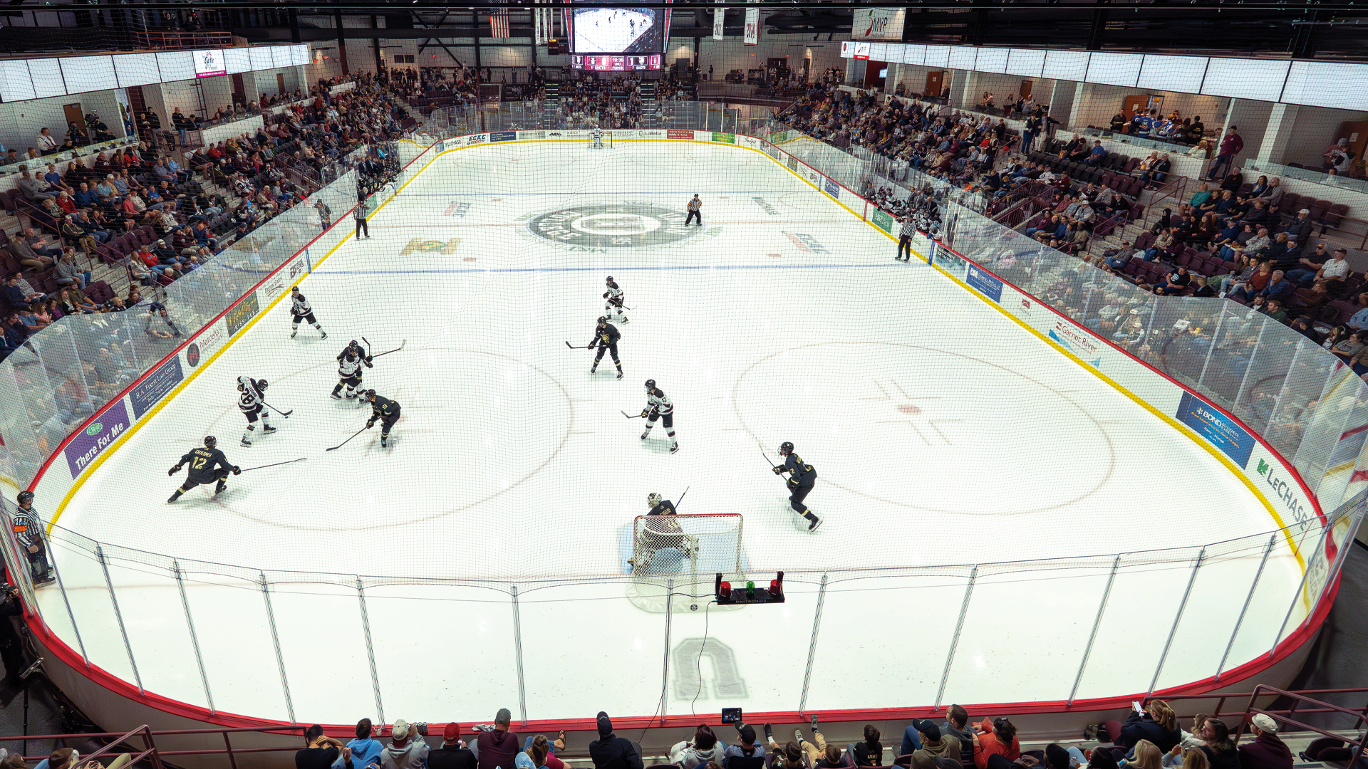 an arial view of a hockey rink from behind the goal with two teams skating on it. One team wears white and the other is in black.