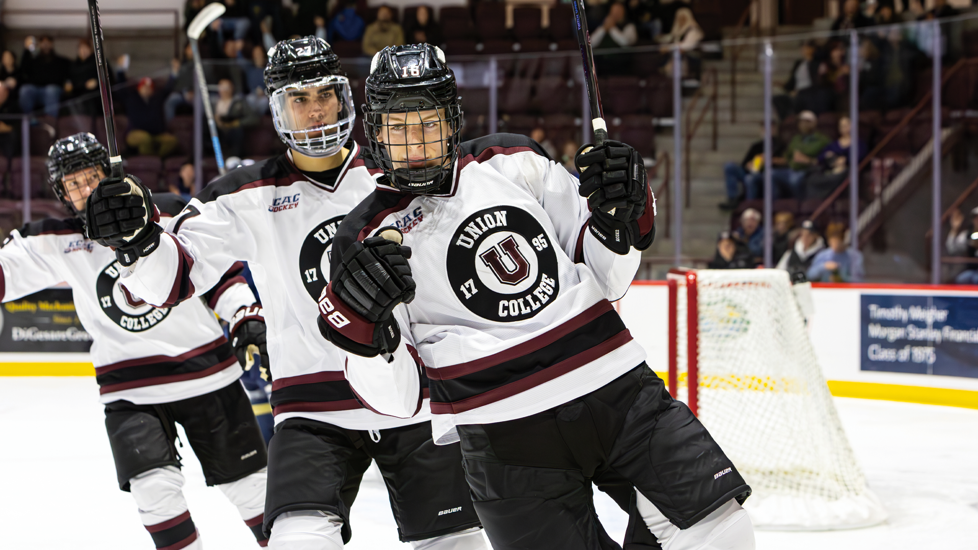 a men's hockey player holding both fists up in celebration after scoring a goal, with two teammates skating behind him also holding their hands up in celebration