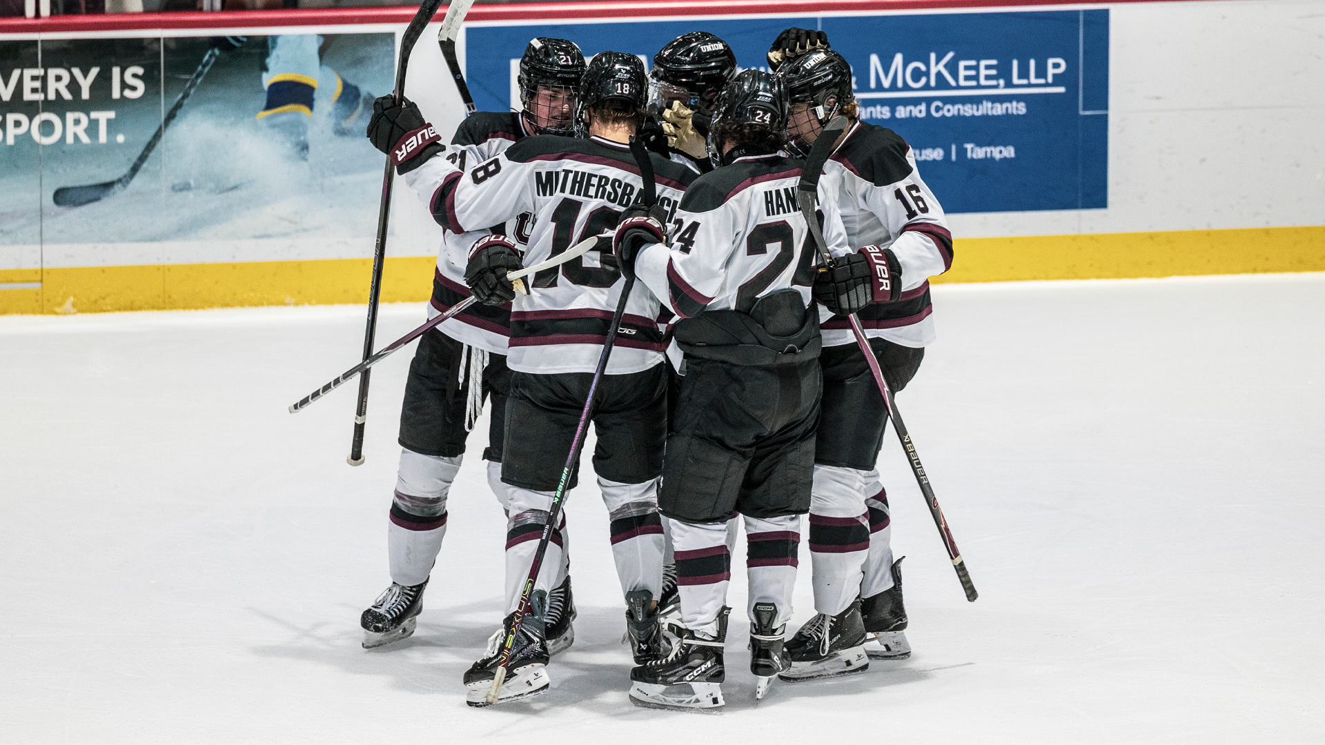 a men's hockey team huddle dup after a goal, wearing white jerseys. Most players have their backs to the camera.