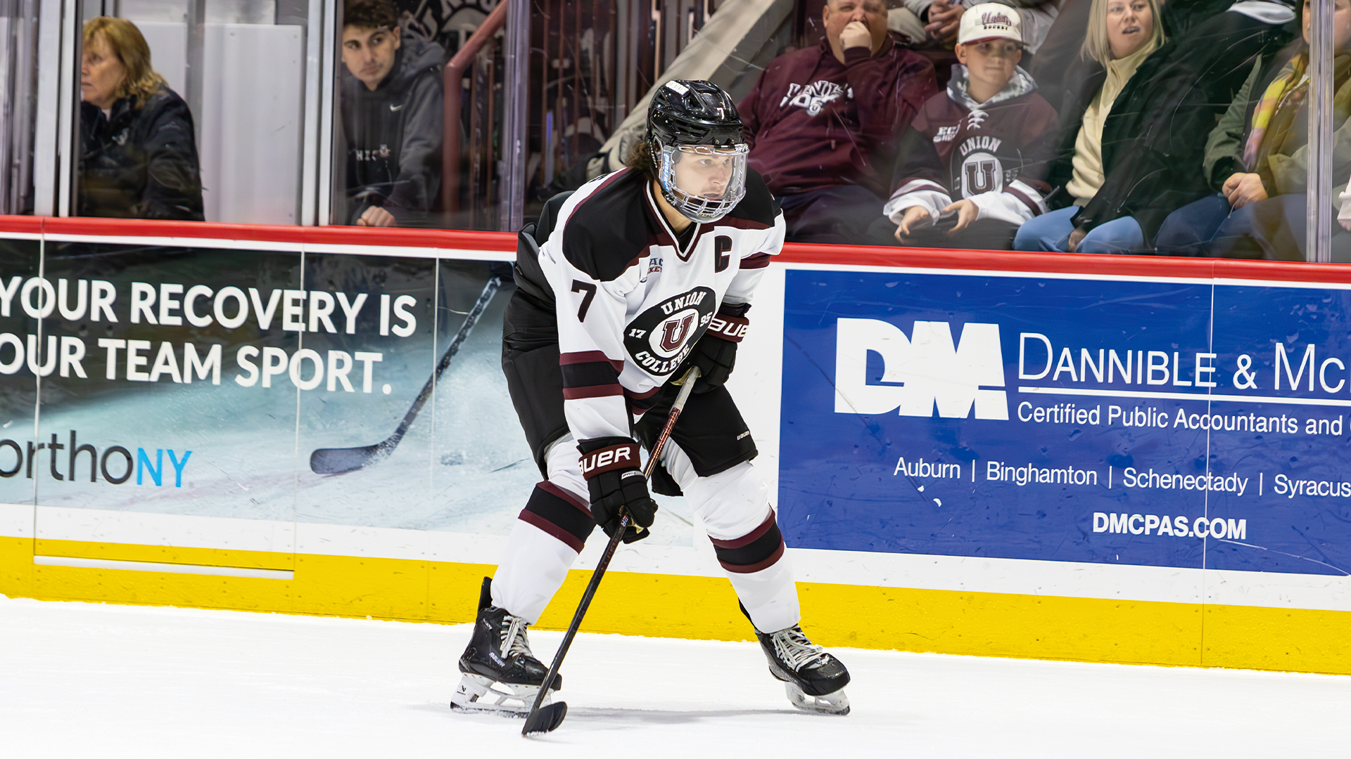 a men's hockey player wearing a white jersey leaning on his hockey stick, out in front of his body