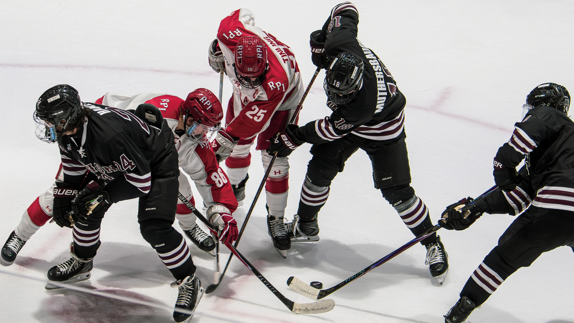 five men's hockey players in the photo, with three wearing black jerseys and two wearing white & red jerseys. Four of the players on the right of the image are looking down and swinging their hockey sticks at the puck, while the fifth player, wearing black, has his back to the play