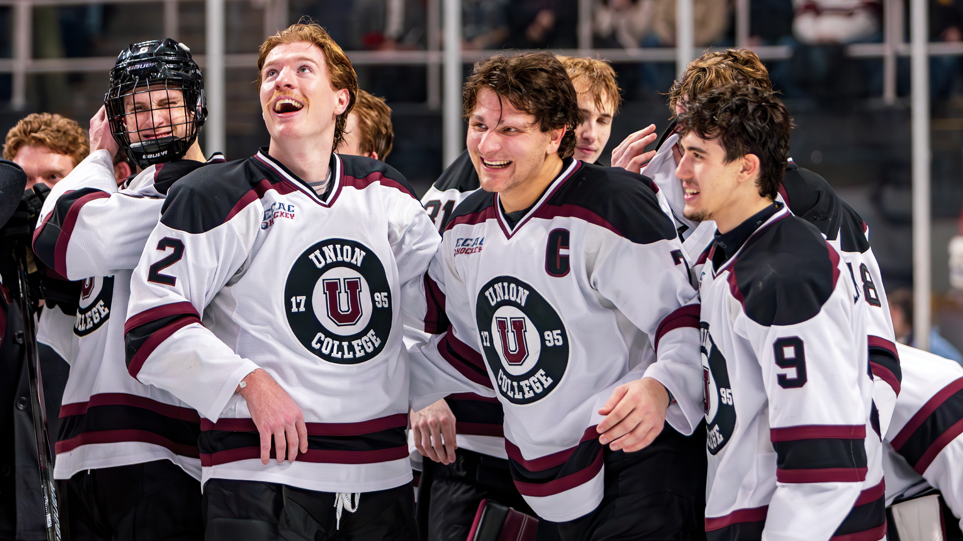 a group of men's hockey players standing on the ice after a big win, with the three players in the foreground of the picture smiling very wide and one player is looking up at the videoboard