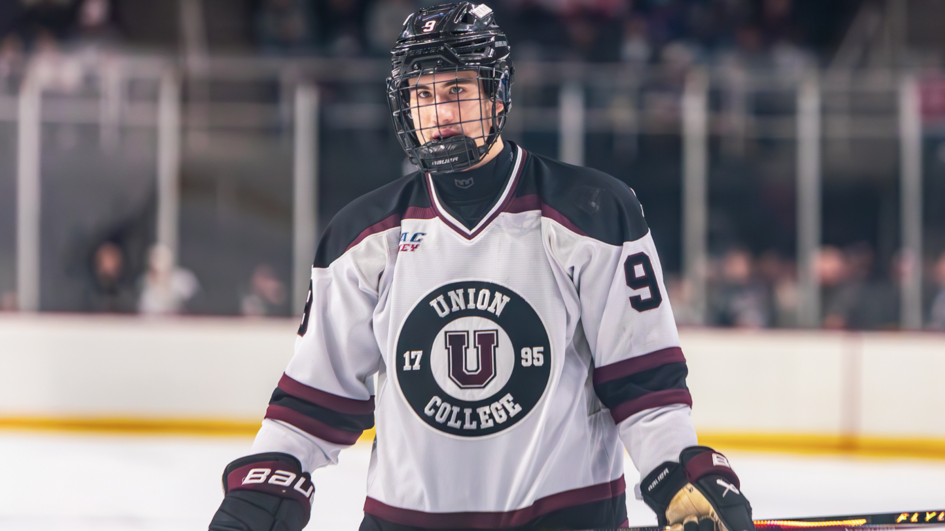 a men's hockey player wearing a white jersey and black helmet, holding his hockey stick across his waist