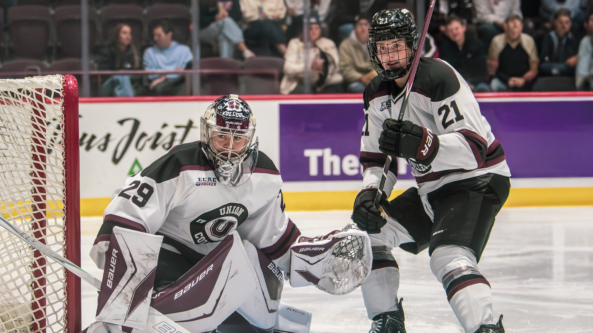 two men's hockey players looking to the left of the frame, with he goalie crouched in front of the near post of the goal and a defender standing to the goalie's left, holding his hockey stick up in the air  
