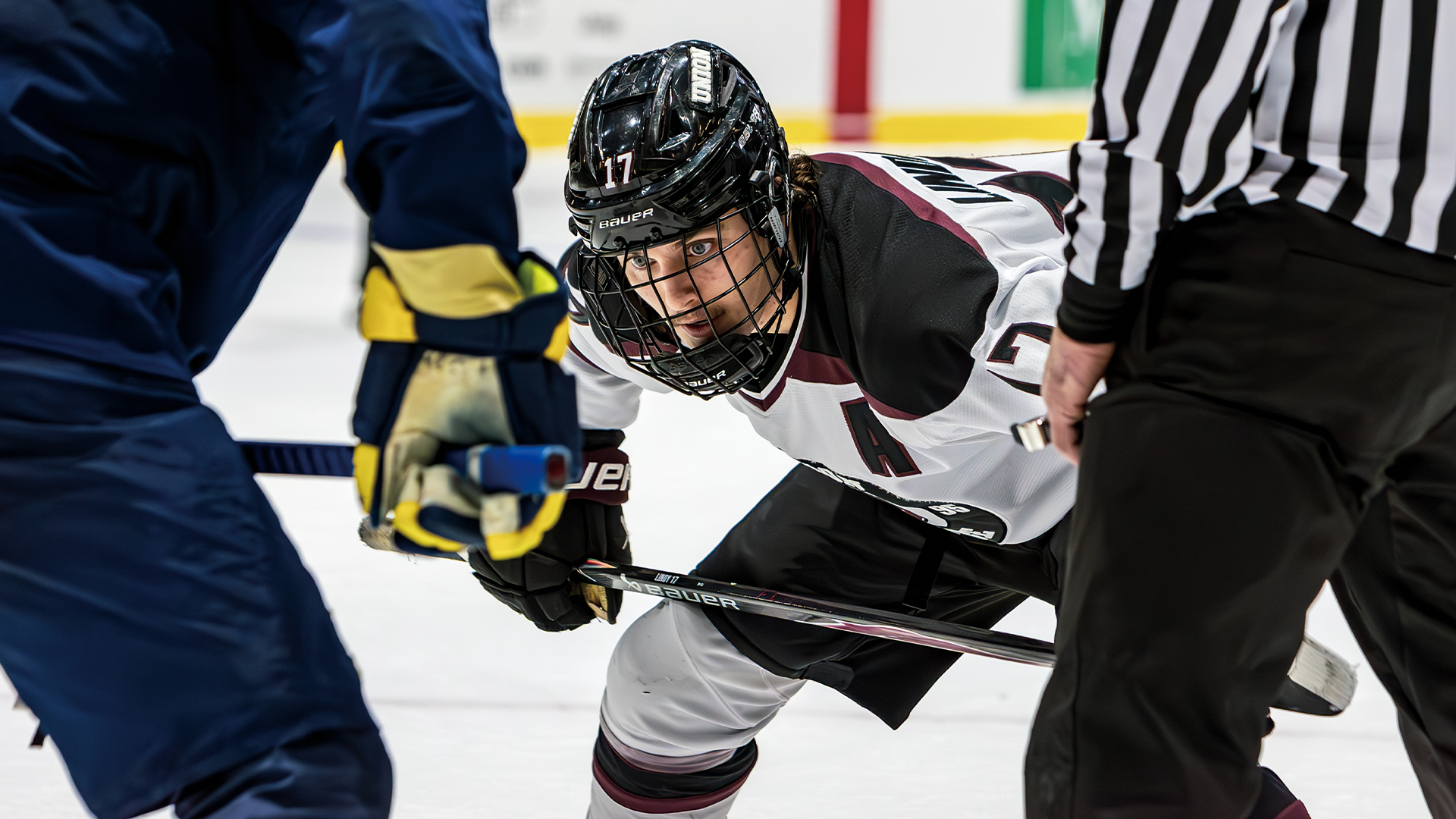 men's hockey player lined up to take a faceoff with a referee to his left and his opponent in front of him. The hockey player is looking intensely at the faceoff dot, which is out of the frame
