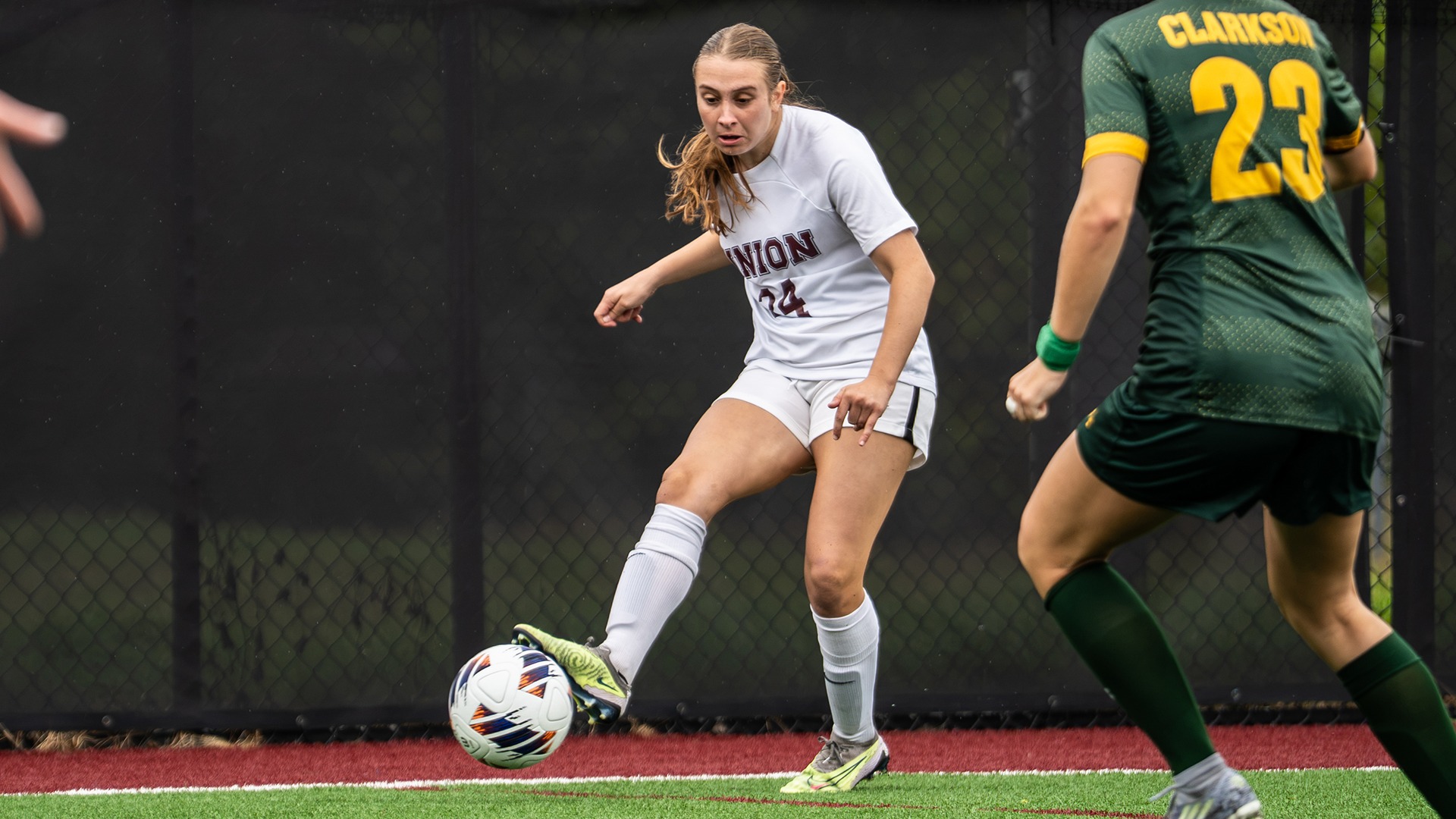 A women's soccer player kicks a ball with an opponent in the foreground.