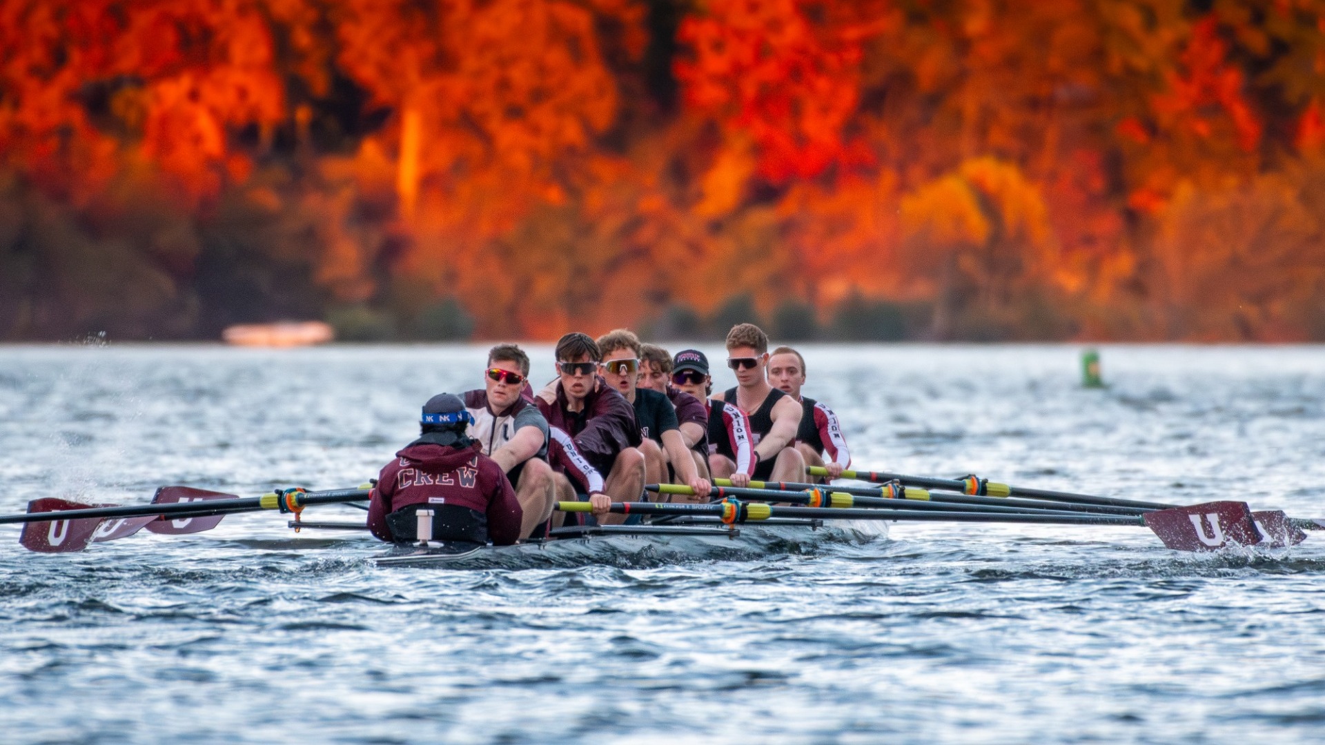 a men's rowing Varsity 8+ boat rowing on rough water with fall foliage in the background