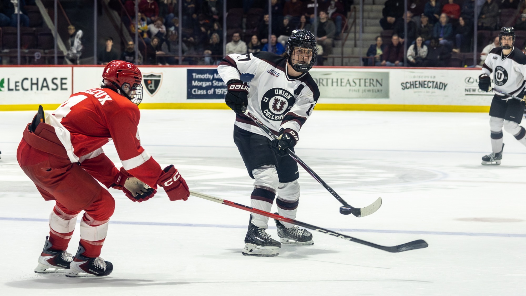 a men's hockey player shooting a puck with a defender wearing all red to his right