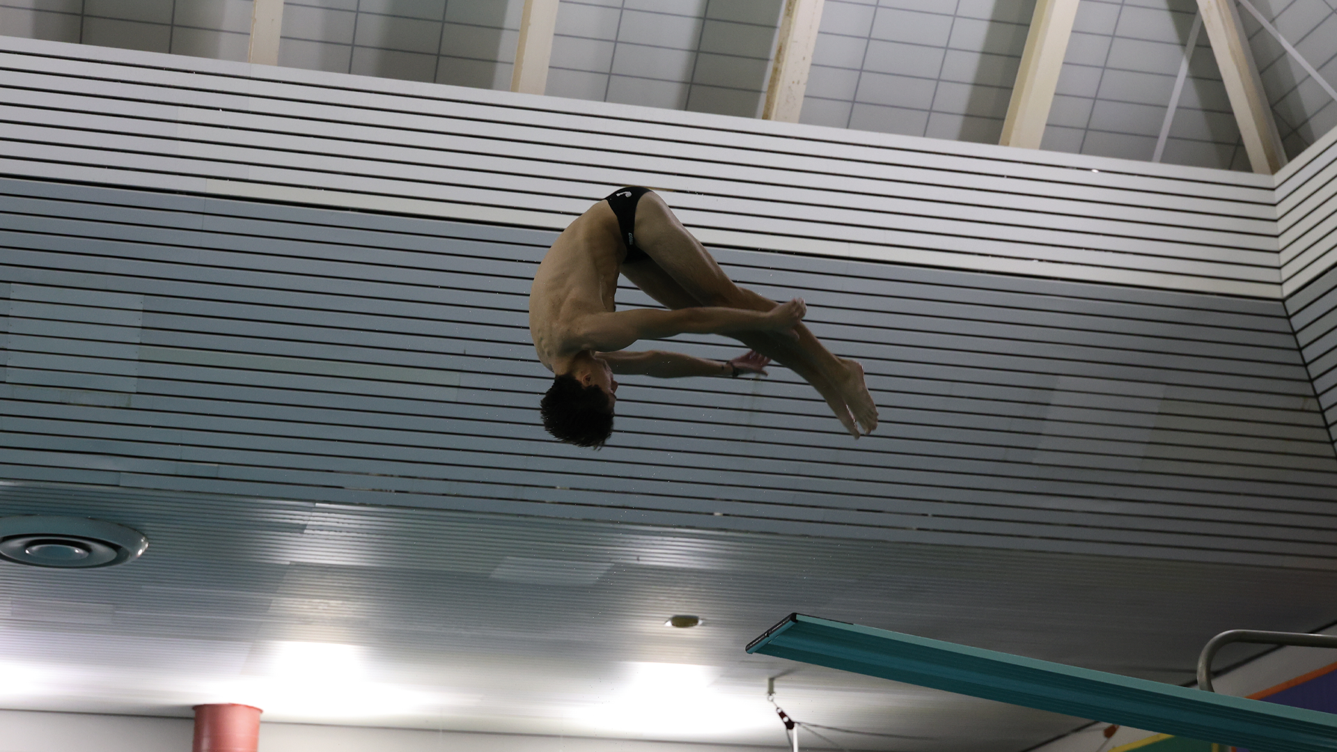 men's diver in the middle of a dive, upside down with his legs at a 90-degree angle to his body. The image is shot from below the diver, looking up at him with the turquoise diving board in the foreground 