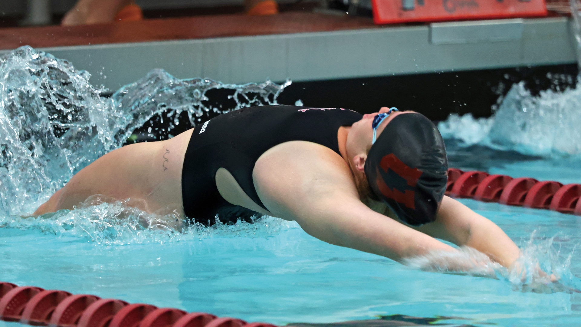 women's swimmer pushing off into the pool to do the backstroke, she is just breaking into the surface of the water, wearing a black suit and black swim cap with a Garnet-colored 