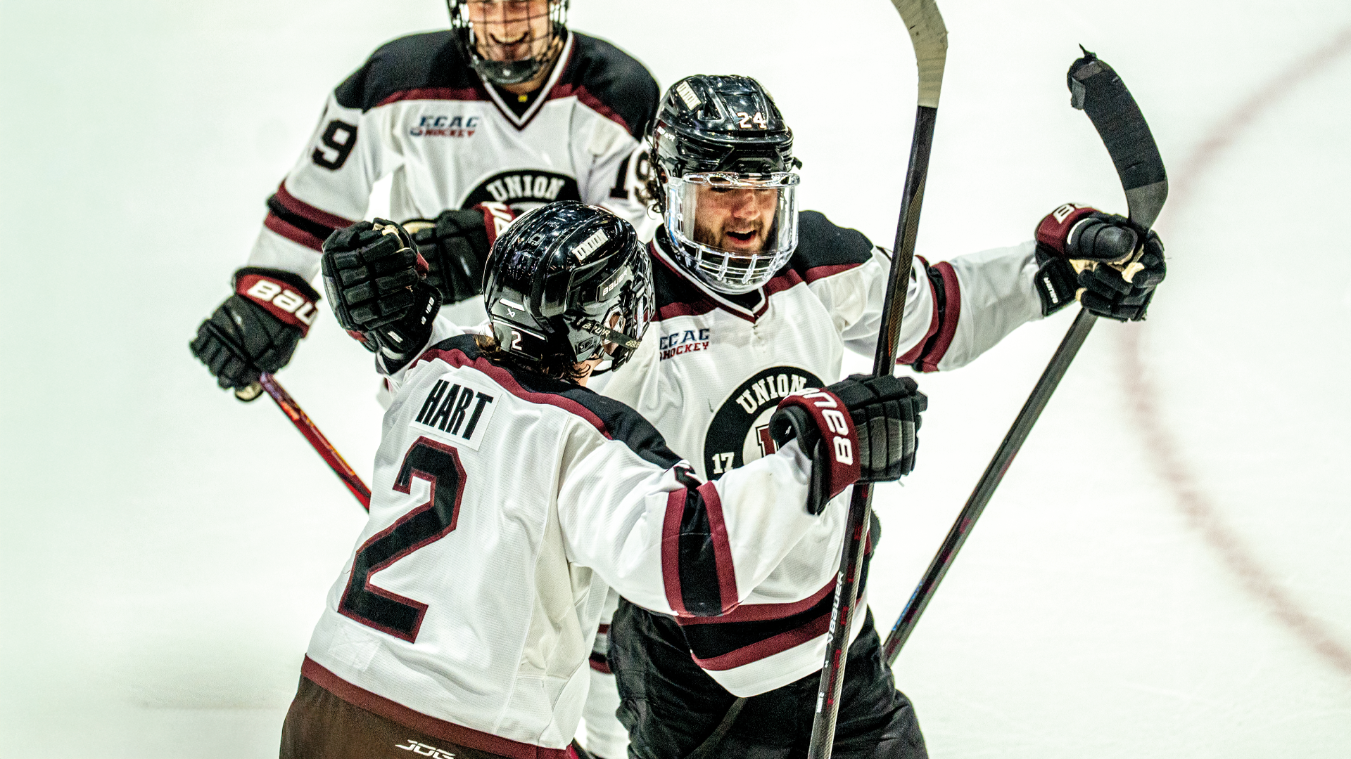three men's hokcey players celebrating along the glass after a goal, with two players about to hug while another skates in from behind them with a smile on his face