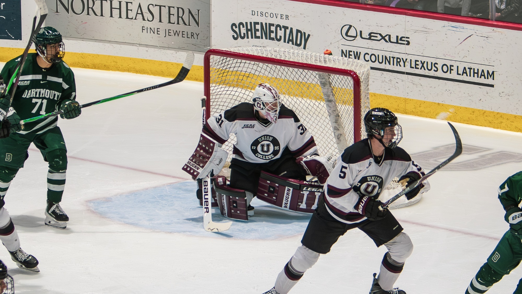 men's hockey goal in his crease looking to his left with an opposing player standing to his right and one of his teammates in front of him