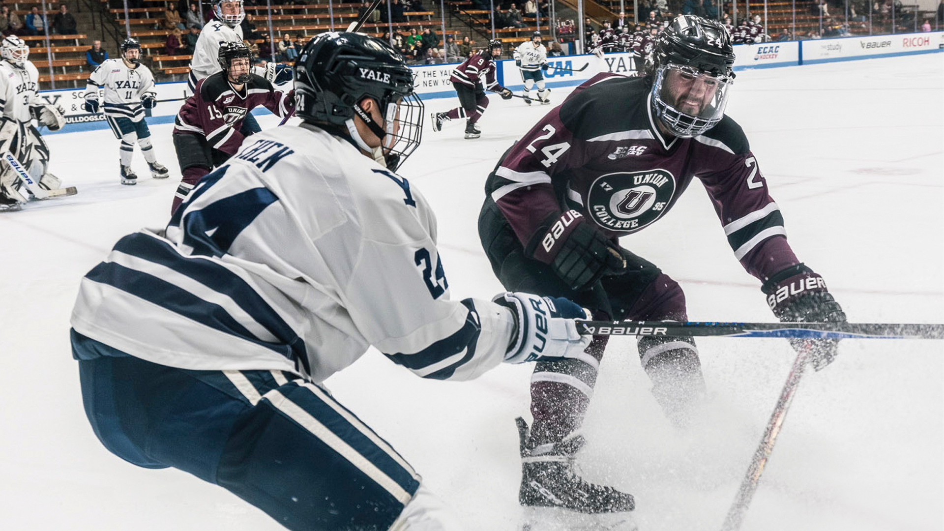 two men's hockey players battling for the puck int he corner, with one player wearing a Garnet-colored jersey and grimacing as he reaches out for the puck, spraying ice in the face of the opposing player wearing white as he stops