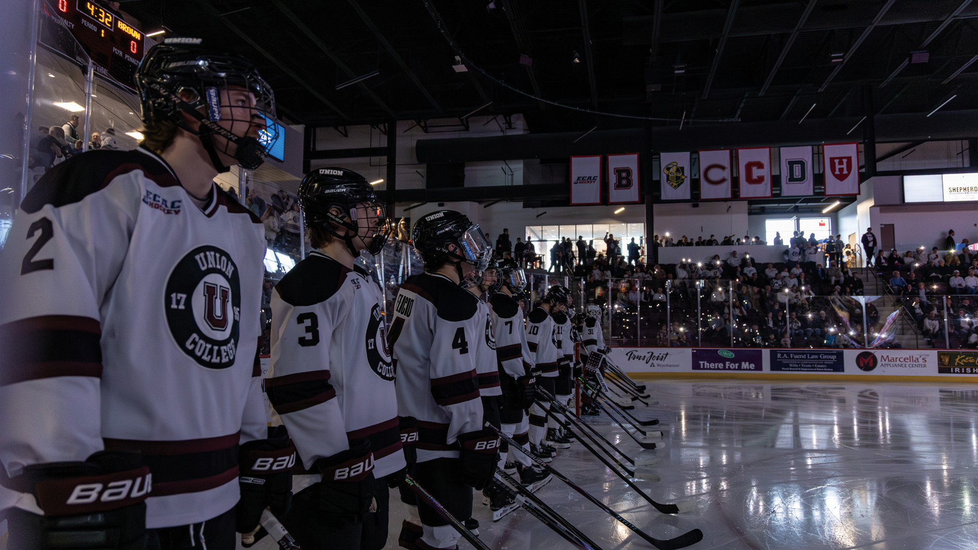 a men's hockey team lined up along the blue line during pregame announcements, with the lights in the arena turned off