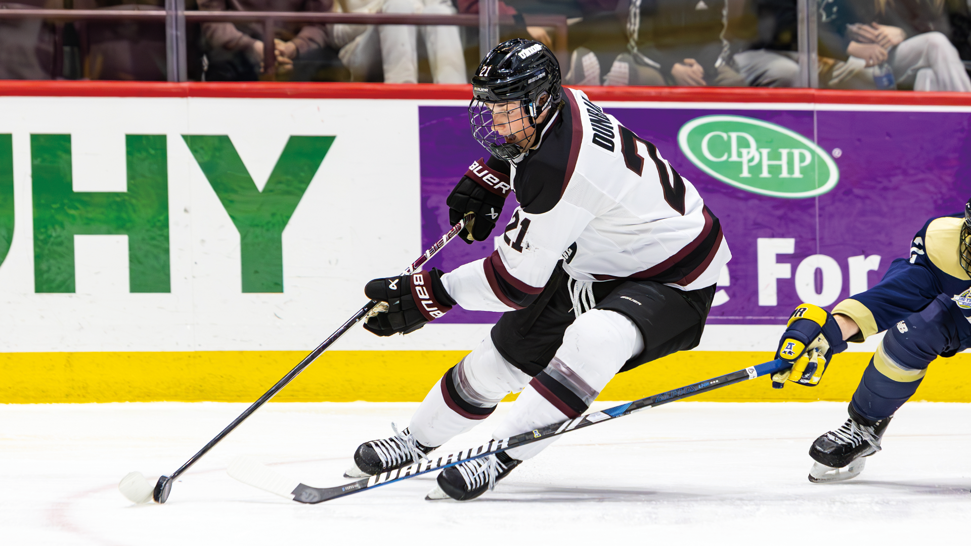 men's hockey player wearing a white jersey and black helmet, handling the puck out in front of him while a defender reaches from behind to try and knock the puck free