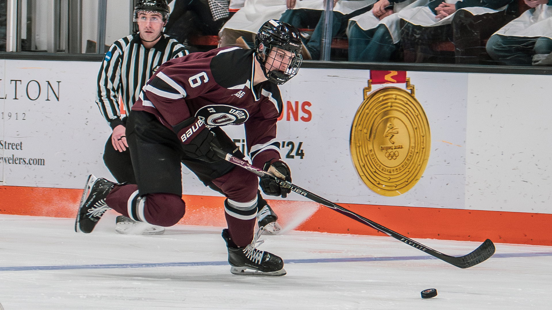 men's hockey player wearing Garnet-colored jersey skating after a puck and looking down at it with a referee behind him