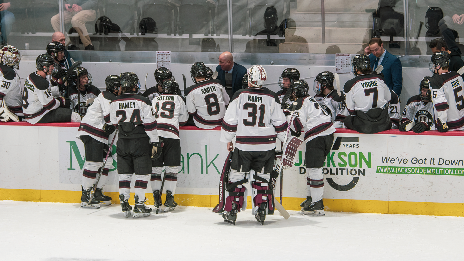 men's hockey team along the boards along their bench with their backs to the camera, listening to their coach speak