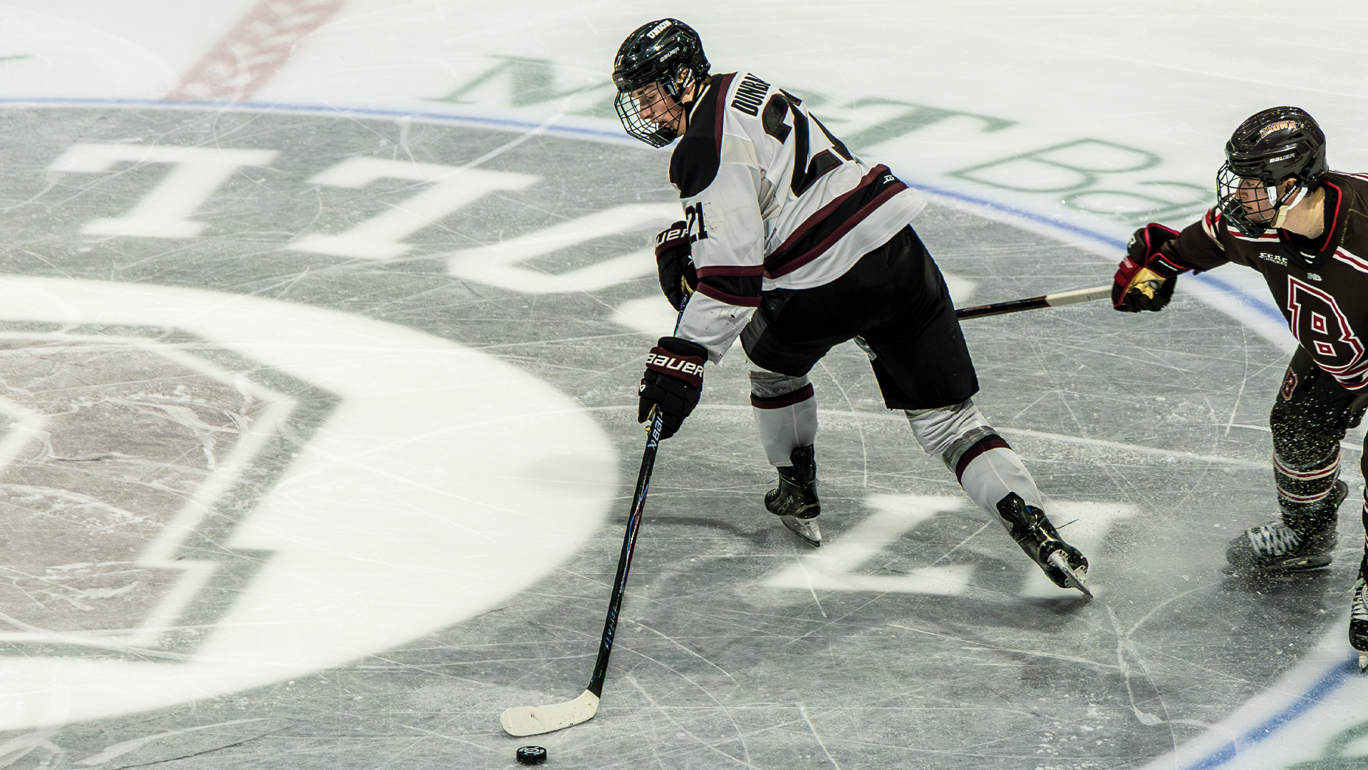 men's hockey player skating across center-ice logo, reaching behind him with his stick to corral the puck while being trailed by a defender