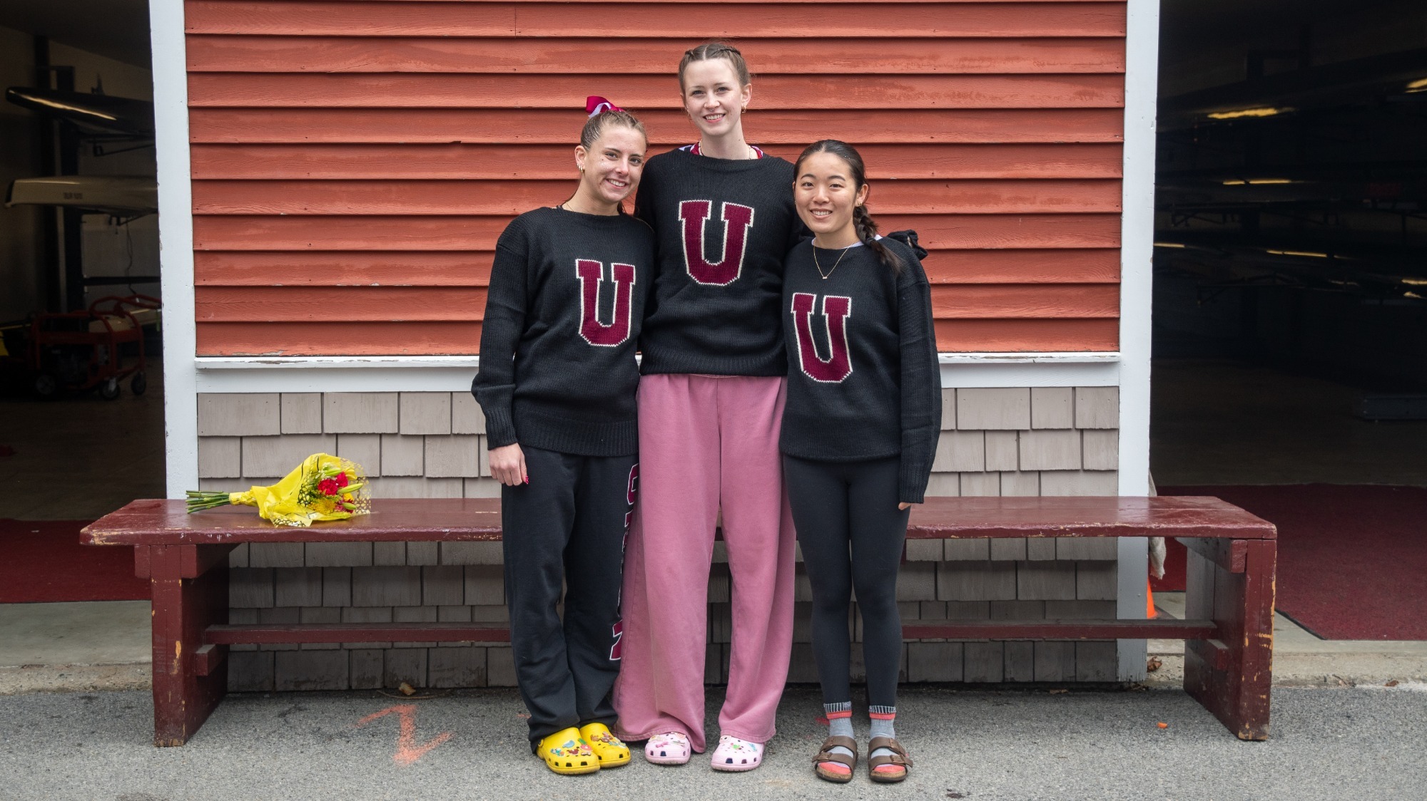 three women's crew athletes standing arm-in-arm smiling at the camera wearing black sweaters with a big block U on the front